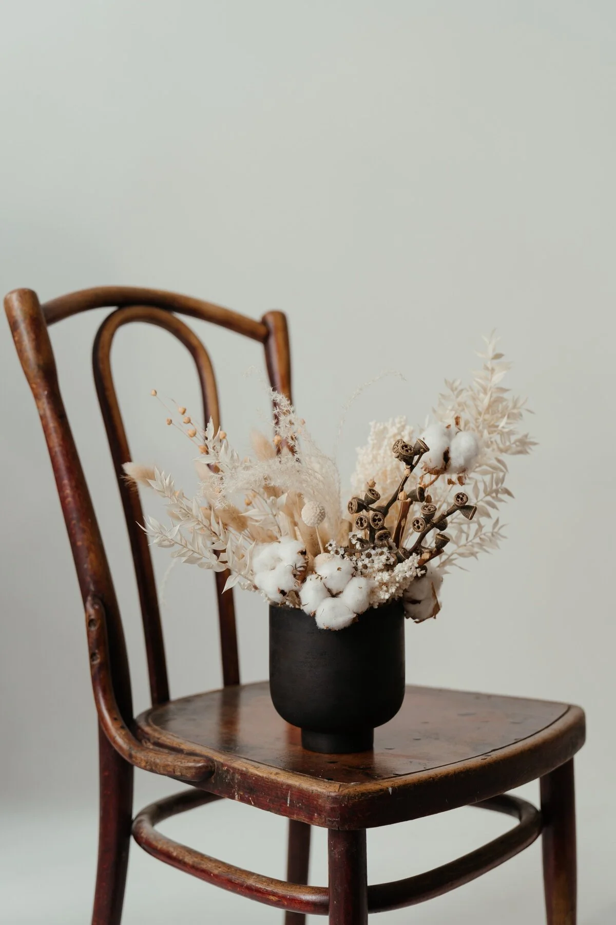 A black vase with dried white flowers and seed pods on a wooden chair.