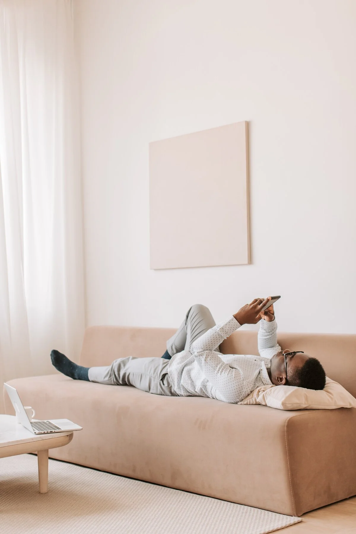 A person lying on a beige sofa, looking at a smartphone, in a minimalist, bright room with a blank wall and white curtains.