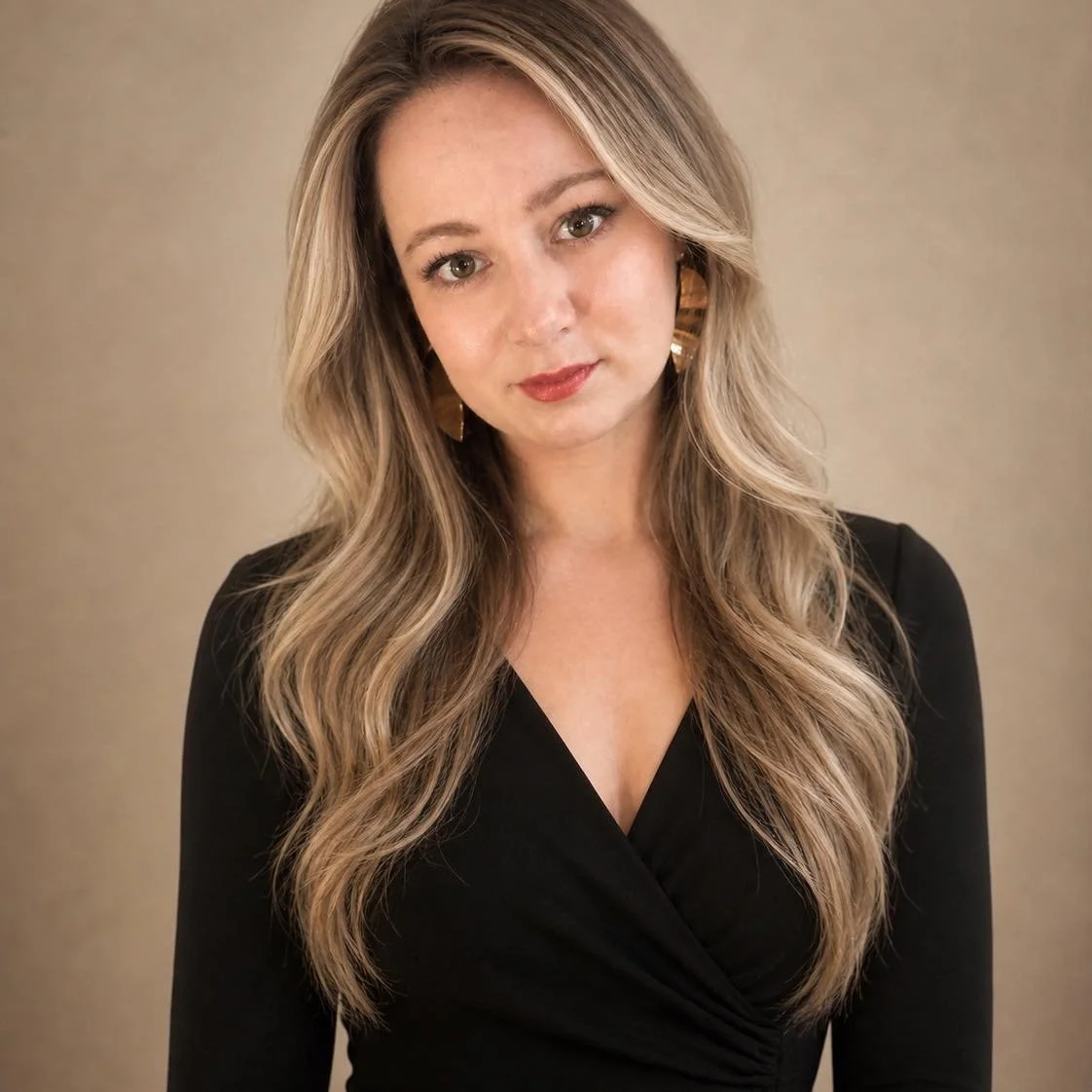 A woman with long, wavy blonde hair wearing a black top with a V-neckline, gold earrings, and subtle makeup, looking directly at the camera against a neutral background.