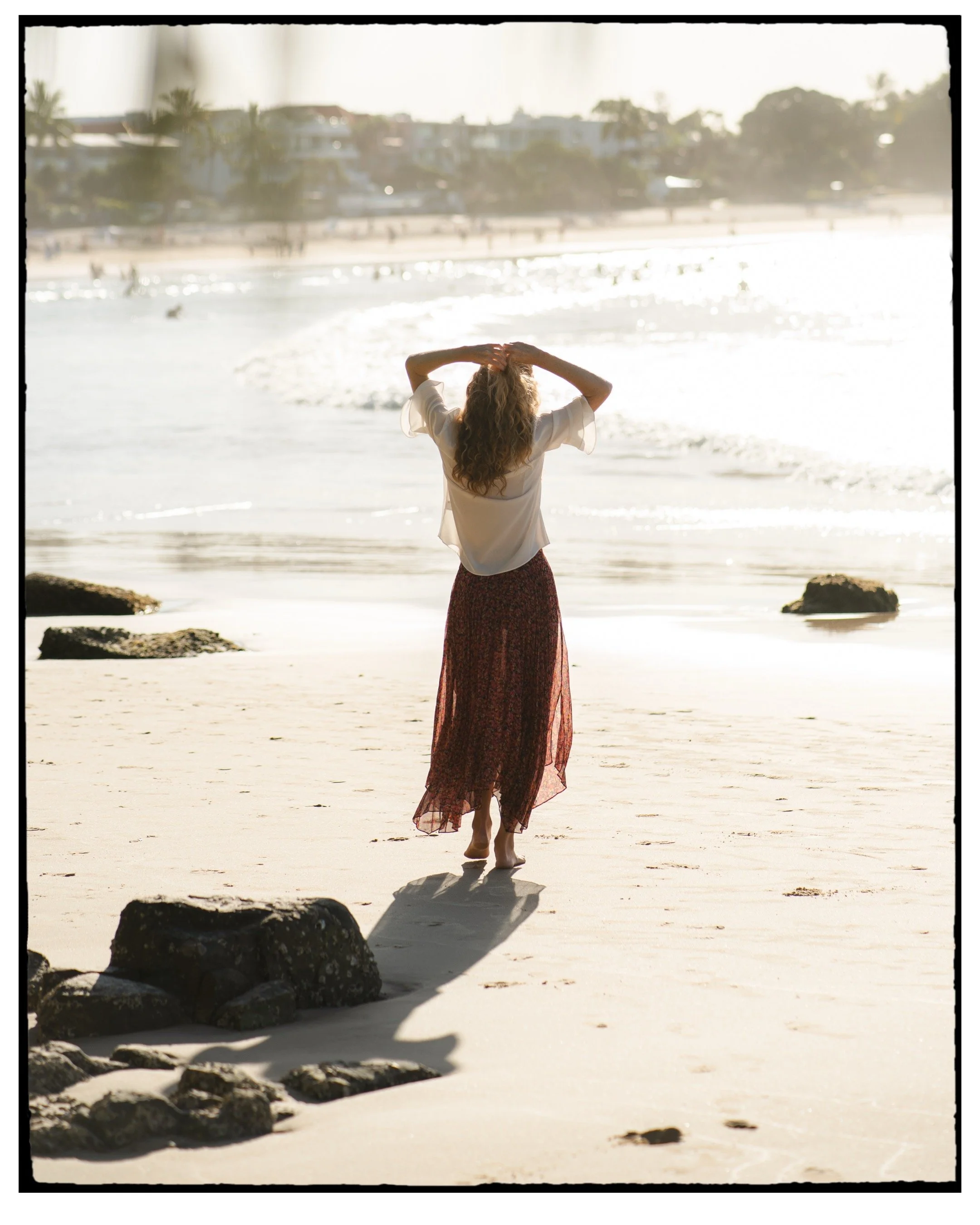 Woman standing on the beach facing the ocean, with her hands on her head, during the daytime with bright sunlight and gentle waves.