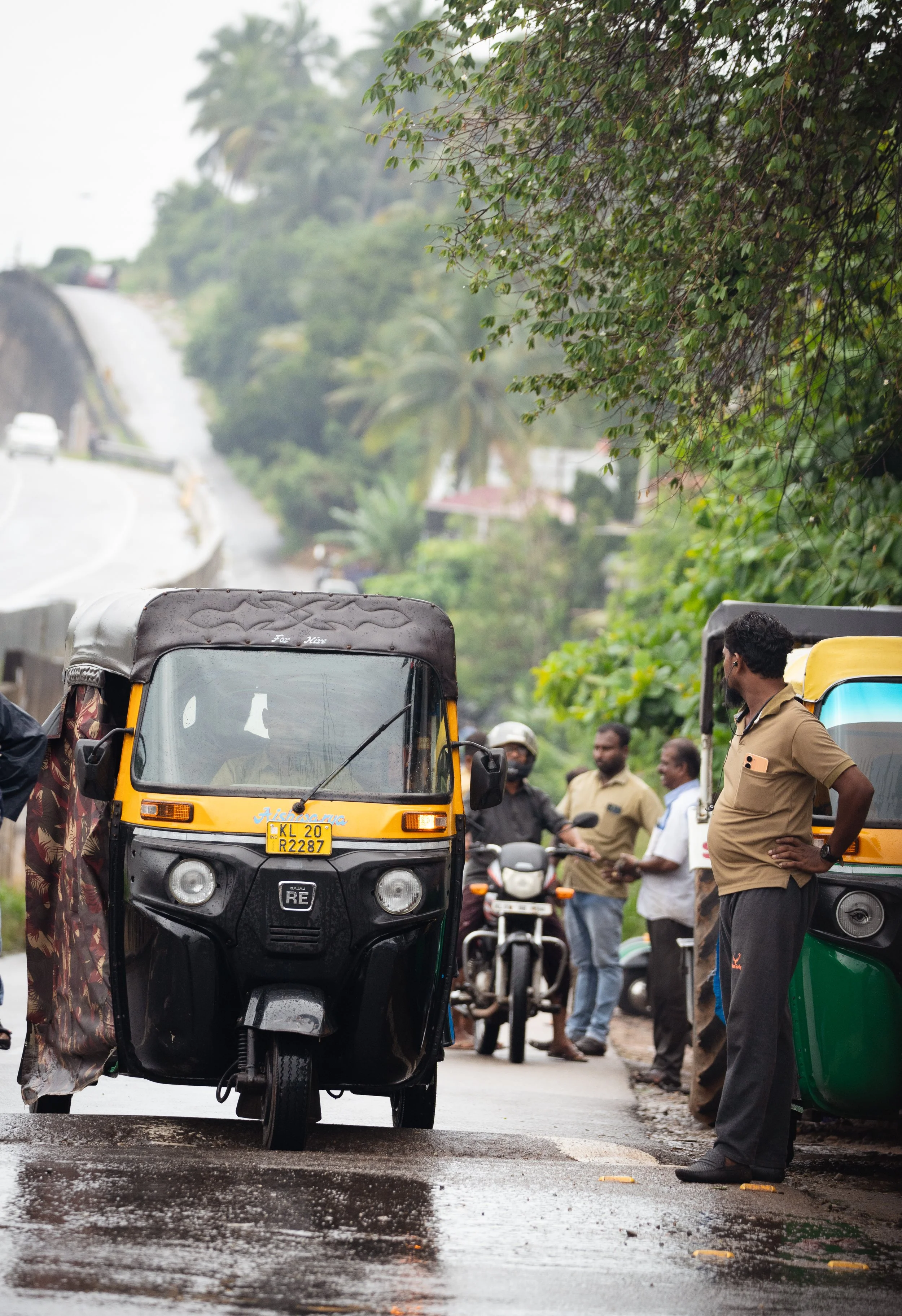 Editorial street photography in India of people gathered beside a rain-soaked road with rickshaws, motorcycles, and a police officer