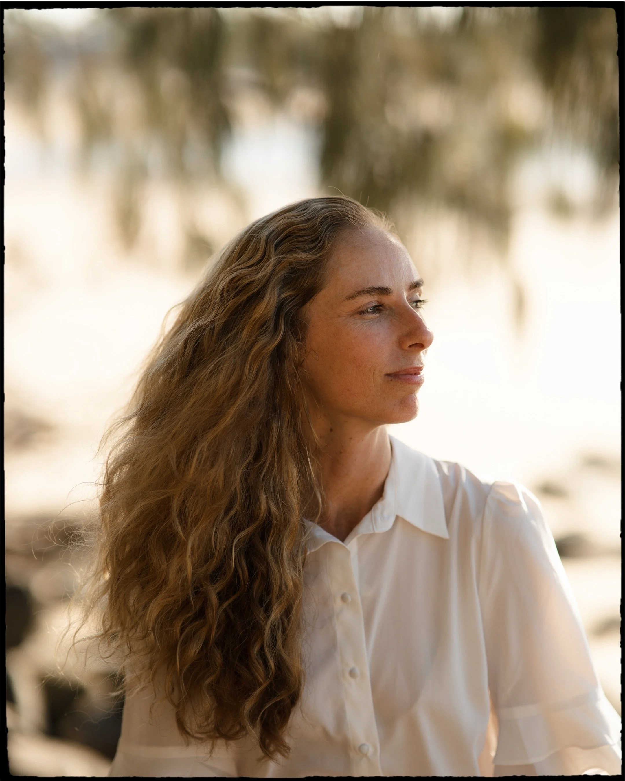 A woman with long, curly brown hair and fair skin wearing a white button-up shirt outdoors with blurred trees in the background.