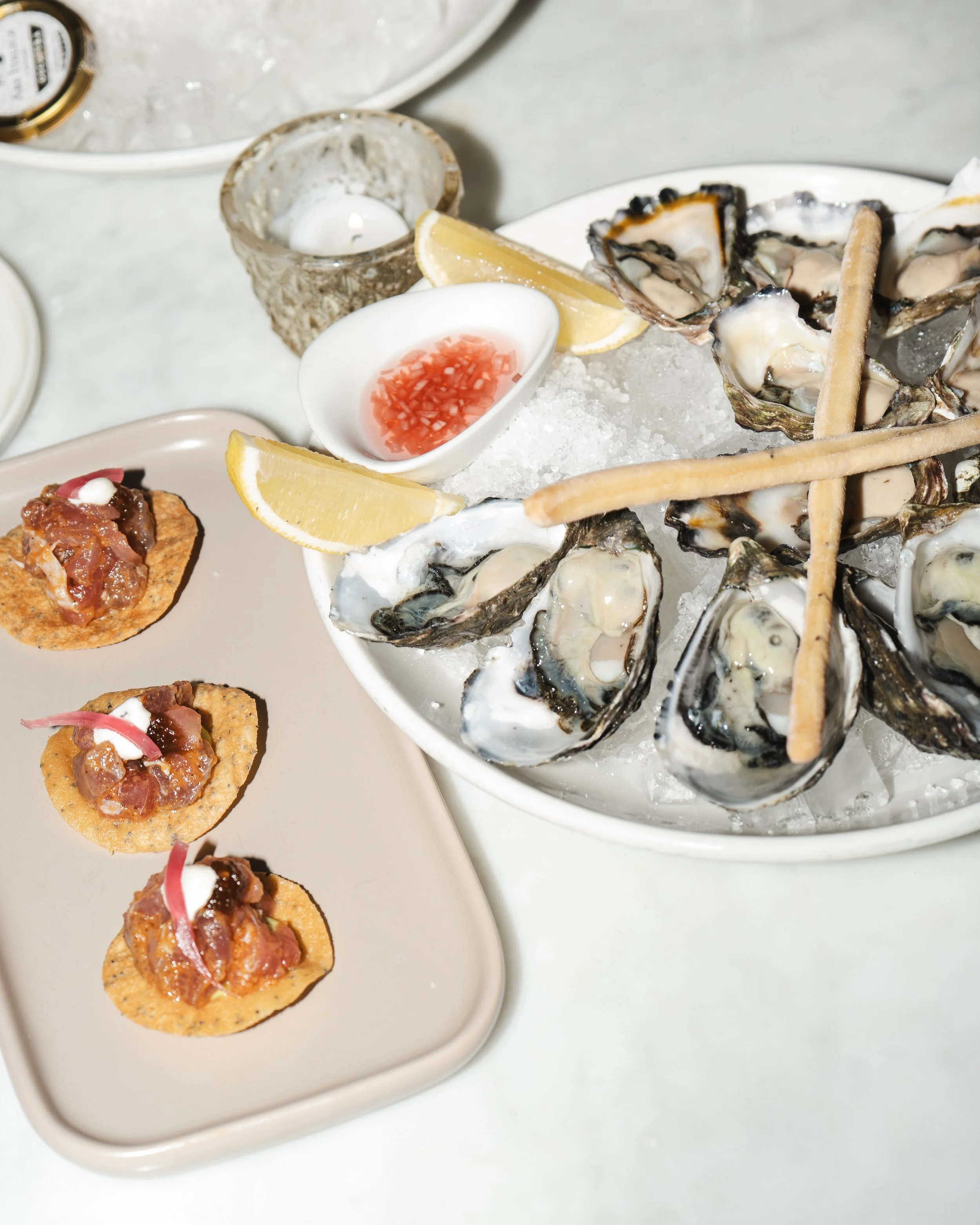 A plate of fresh oysters on ice, garnished with lemon wedges and served with cocktail sauce, next to a small dish of red sauce and a lit candle in a decorative holder, and a smaller plate with three canapés topped with a mixture of chopped ingredient