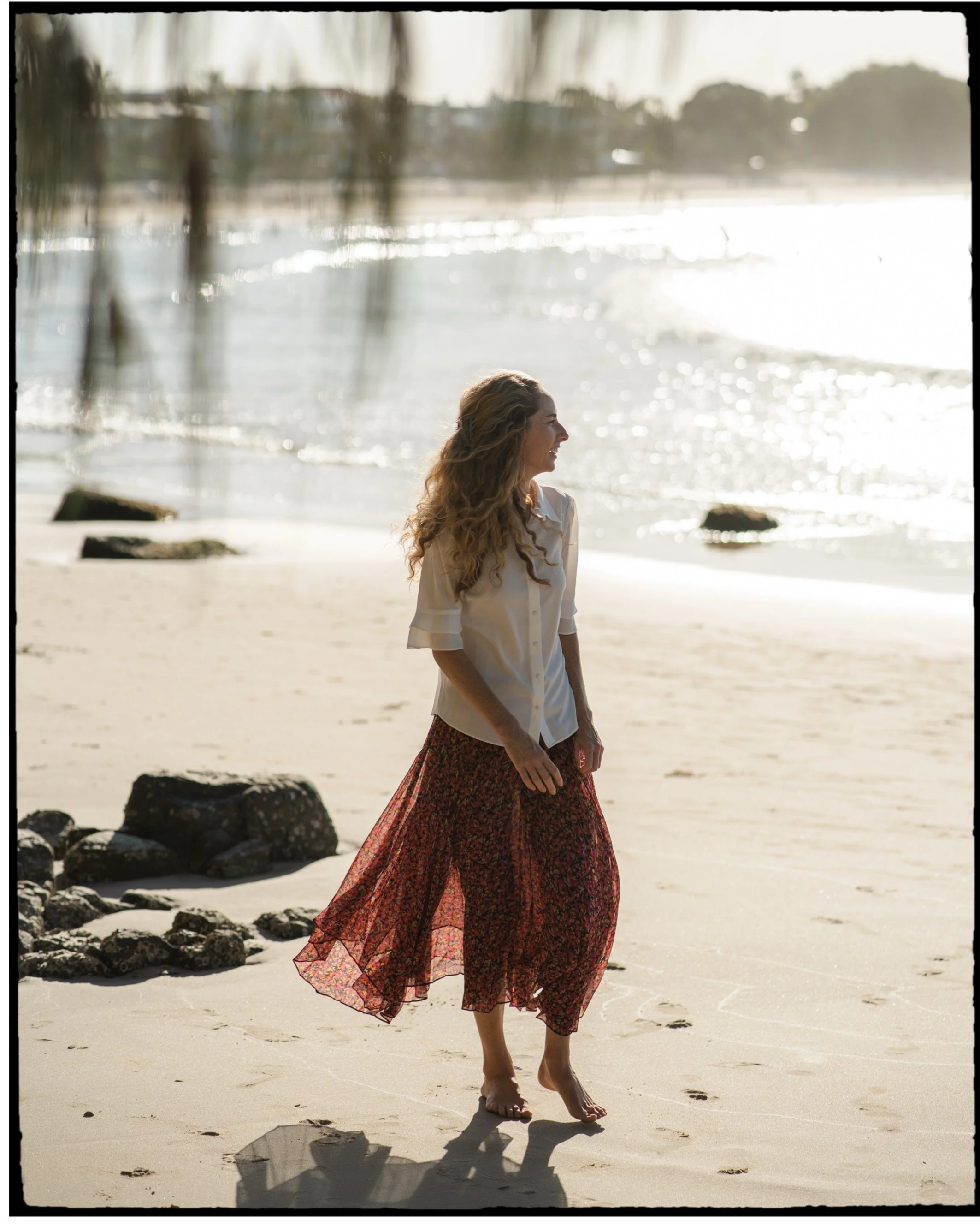 A woman walking barefoot on a sandy beach near the water, wearing a white blouse and a red floral skirt, with trees and the ocean in the background.