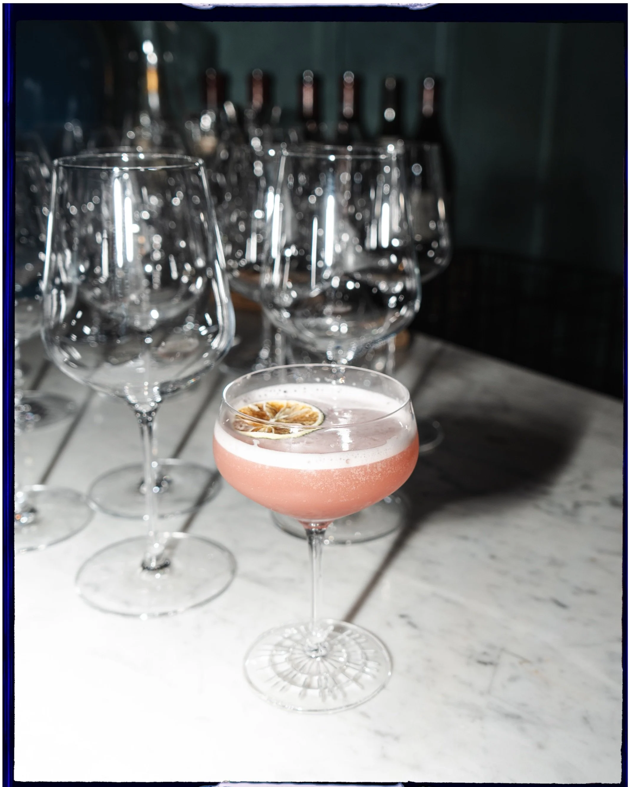 A pink cocktail with a lemon slice garnish in a coupe glass, surrounded by empty wine glasses on a marble bar counter.