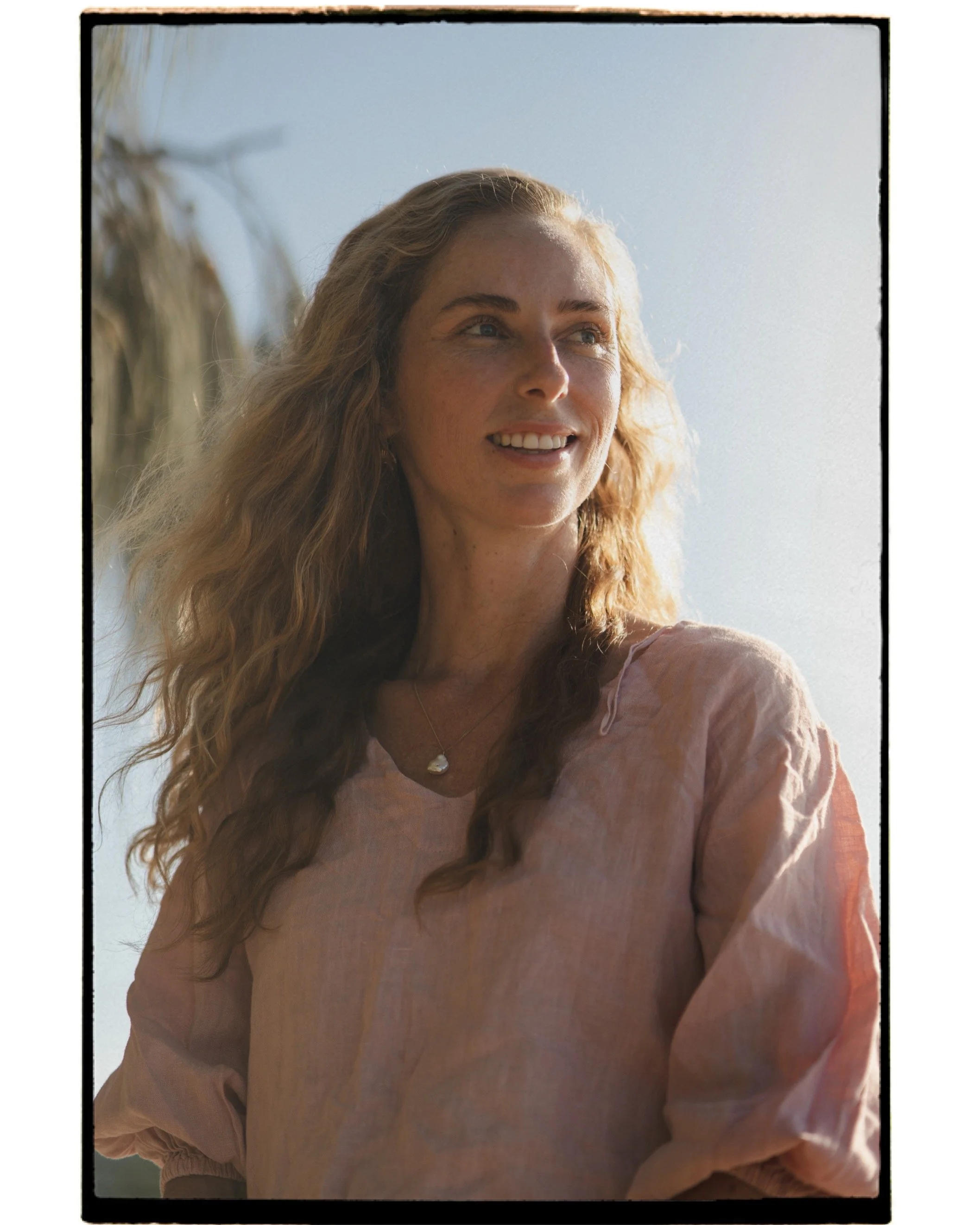 A woman with long, wavy blonde hair smiles while looking off to the side, wearing a light pink top and a necklace with a seashell pendant, against a bright sky background.