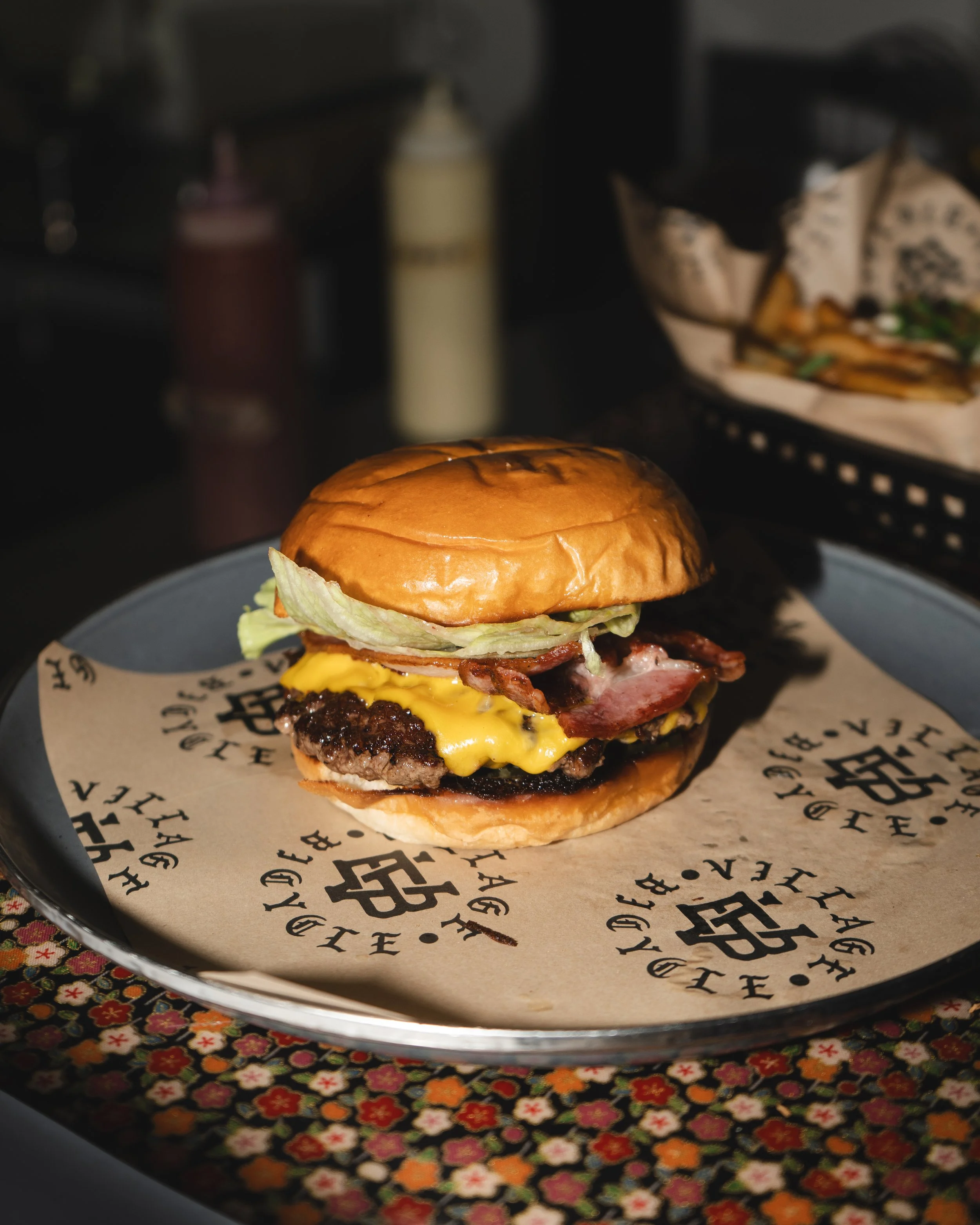 A cheeseburger with bacon, lettuce, and a sesame seed bun on a tray lined with branded paper.