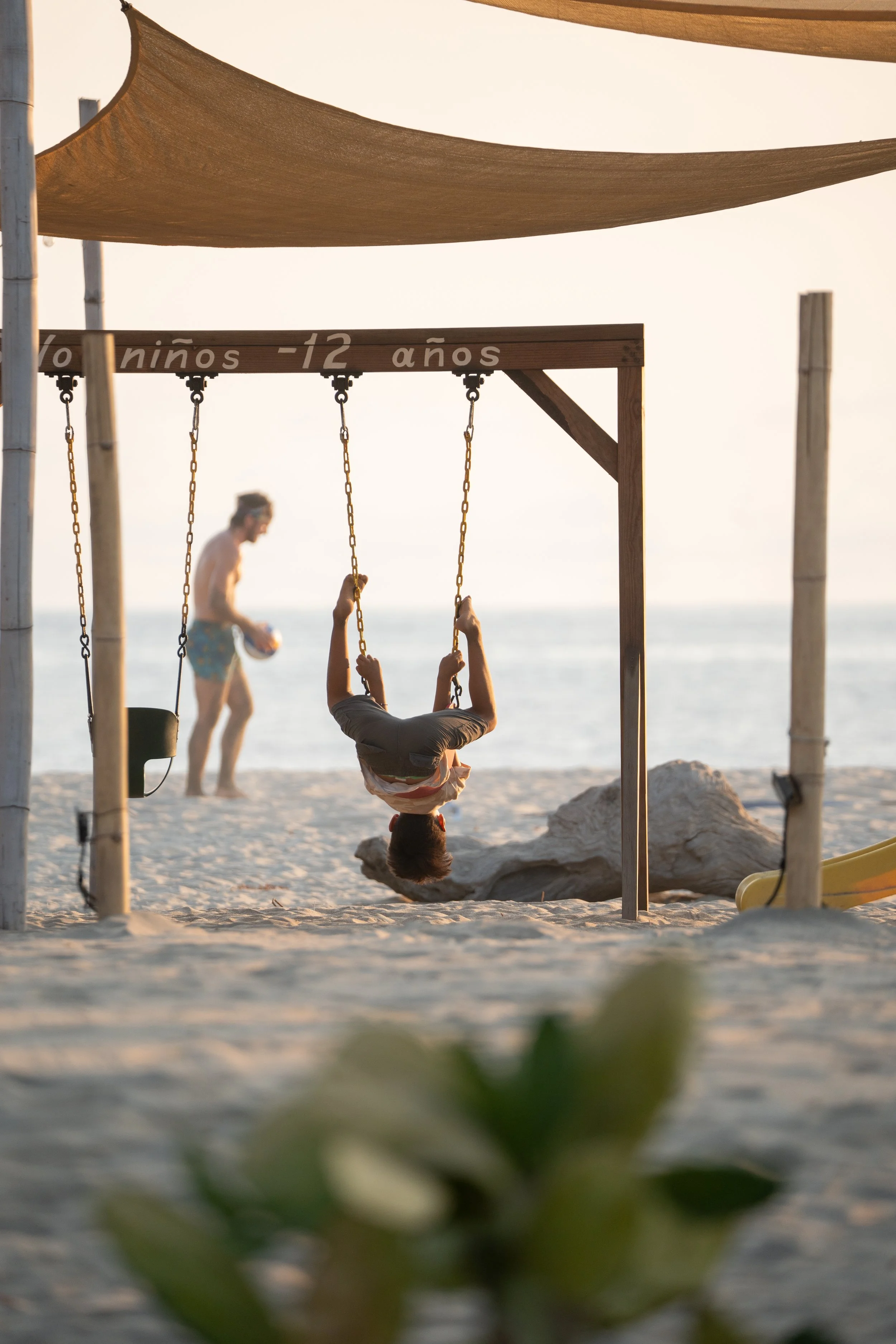 Editorial travel photography of a child playing on a beach swing at sunset with an adult in the background