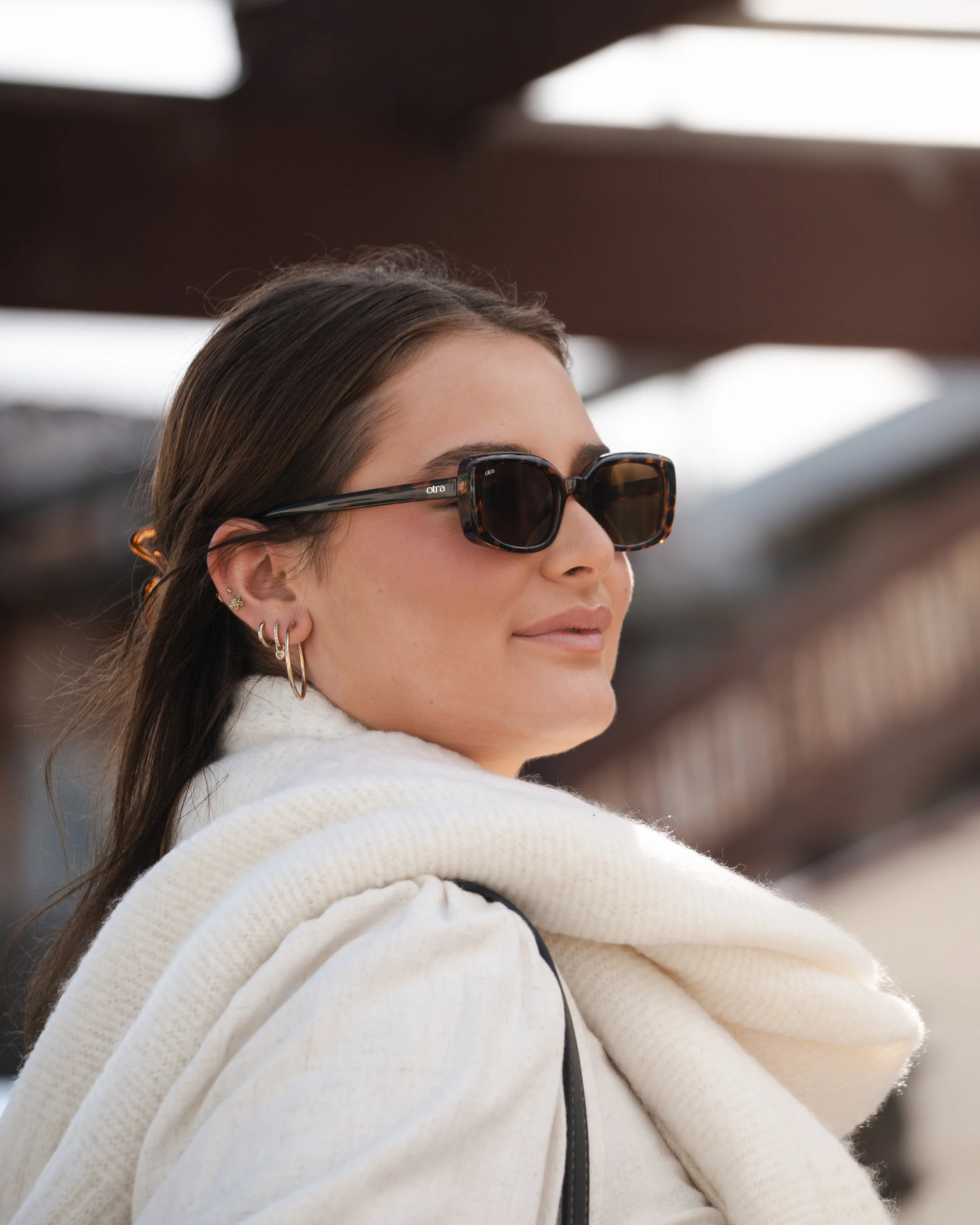 A woman with long dark hair wearing large black sunglasses, earrings, and a white hoodie with a black strap over her shoulder, outdoors.