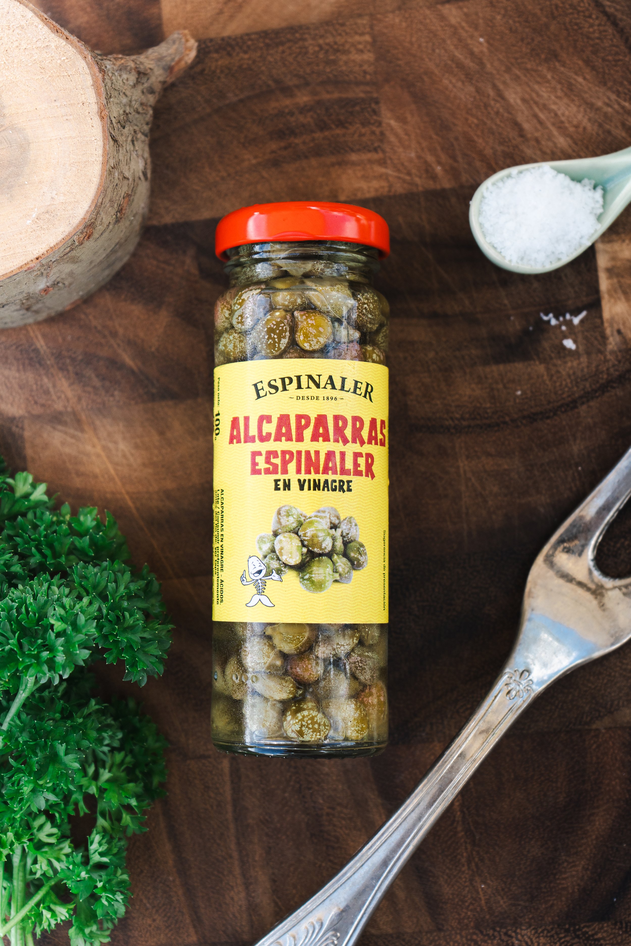 A jar of Alcaparras Espinaler preserved capers on a wooden surface, with a spoonful of salt, a fork, and some herbs nearby.