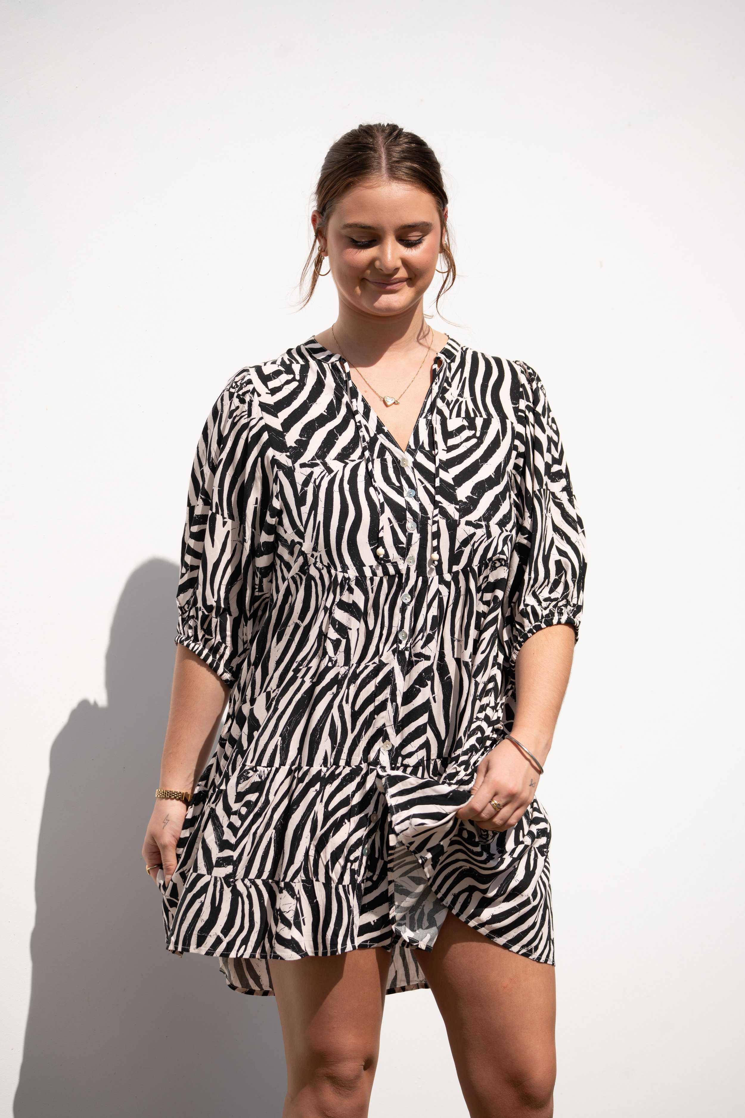 A young woman with brown hair tied back, wearing a black and white zebra print dress, standing against a white background with her eyes closed and smiling gently.