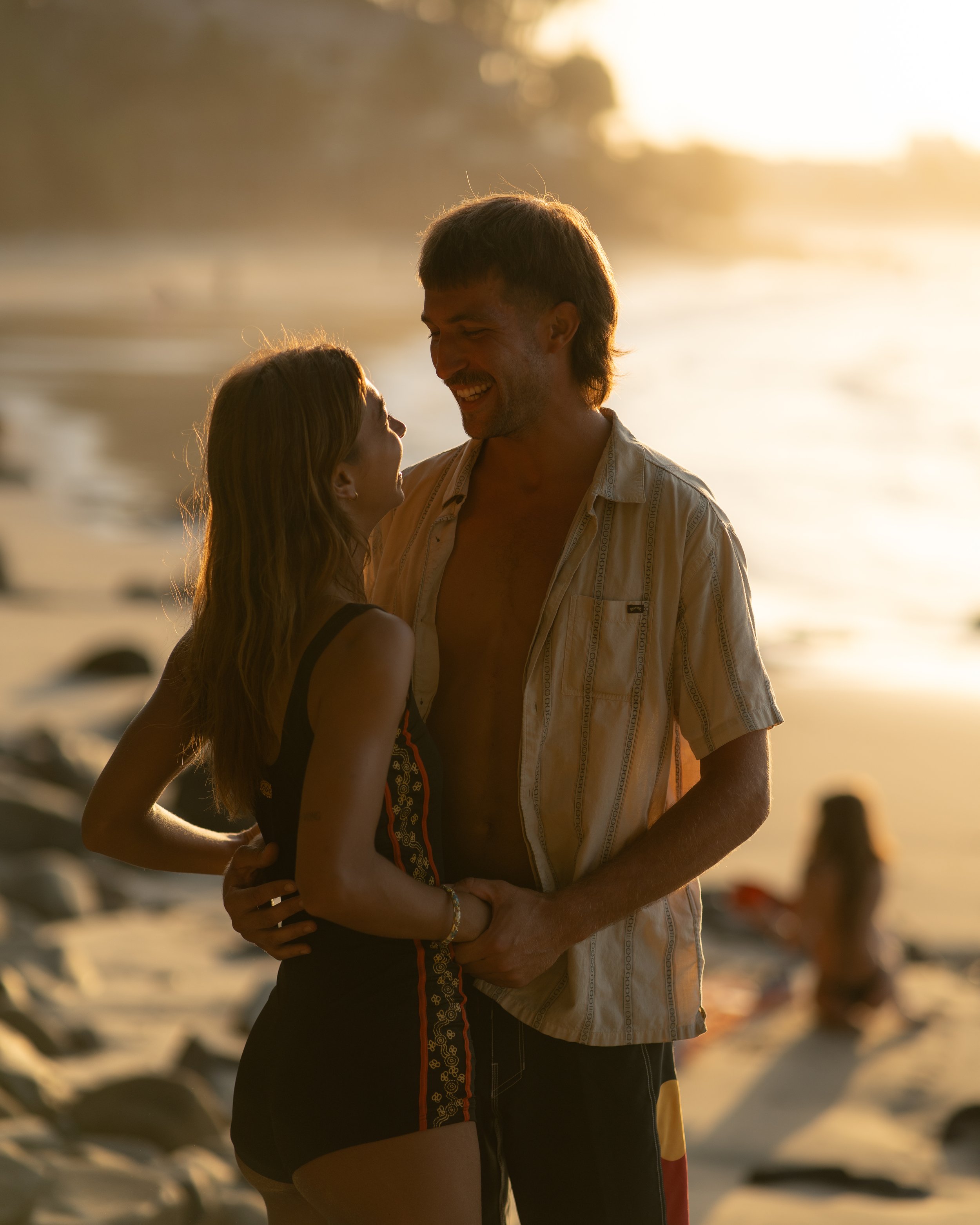 A couple smiling and gazing at each other on a beach during sunset.
