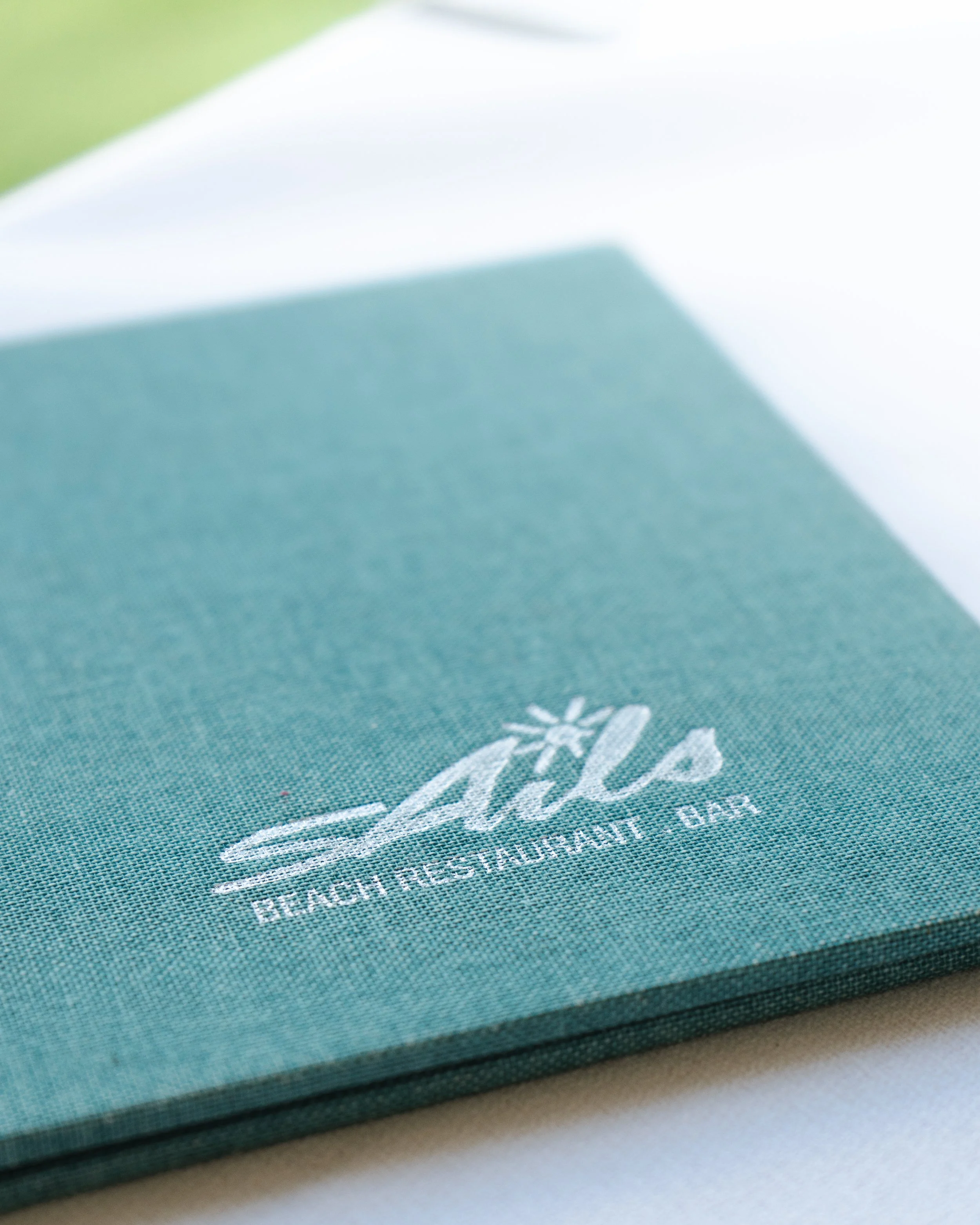 A teal cloth napkin with white embroidered logo reading "Seas" and smaller text "Beach Resort Restaurant Bar".
