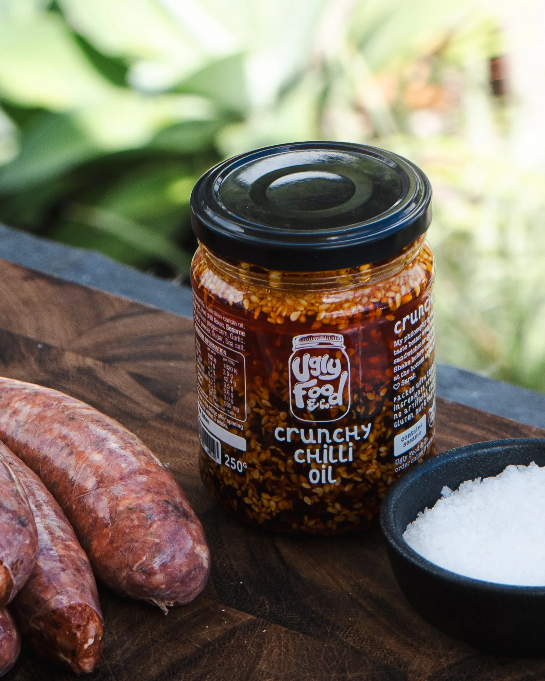 A jar of crunchy chili oil on a wooden surface next to sausages and a small bowl of salt, with a blurred green outdoor background.