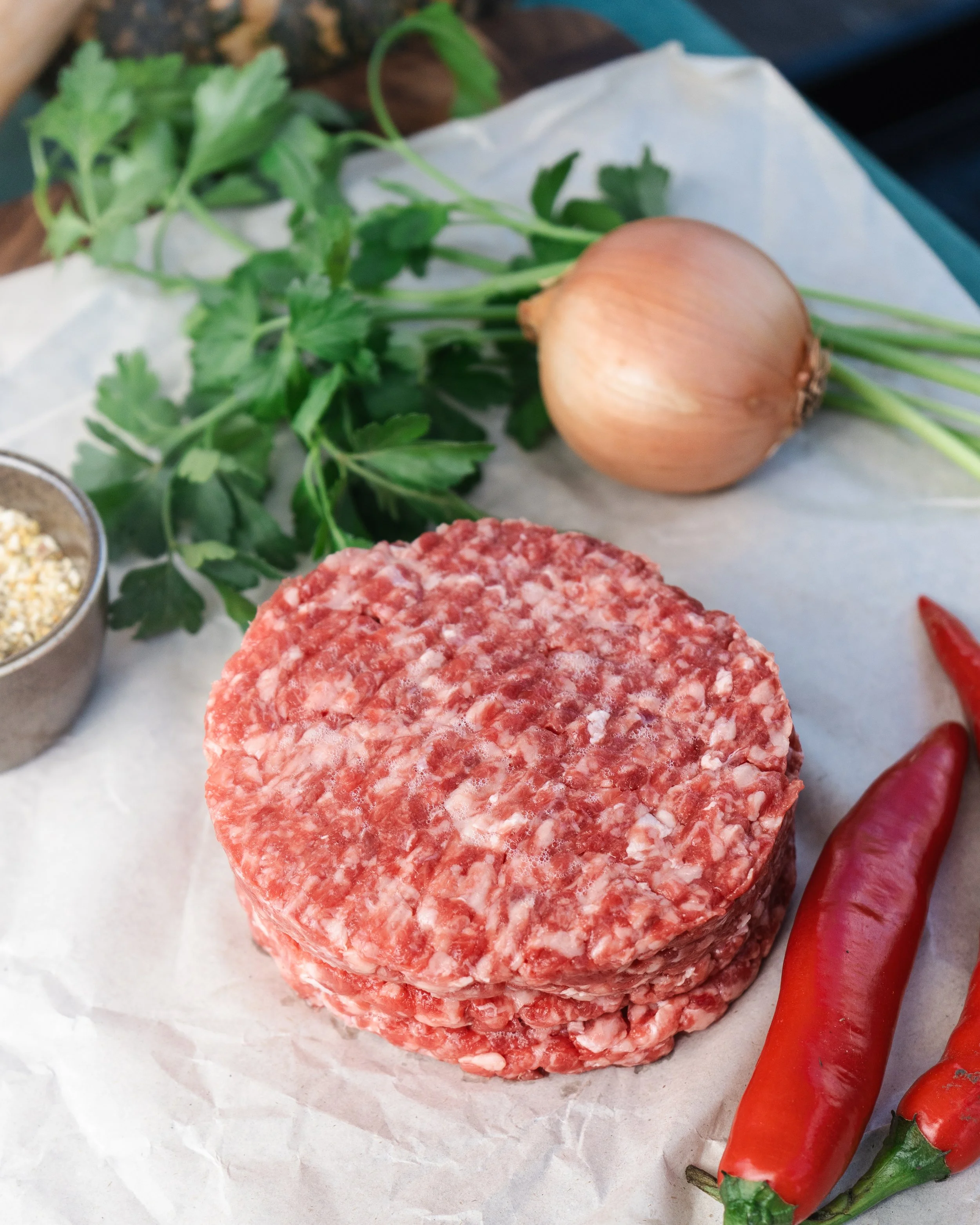 Raw ground beef patties, onion, red chili peppers, fresh parsley, and seasoning in a bowl on parchment paper.