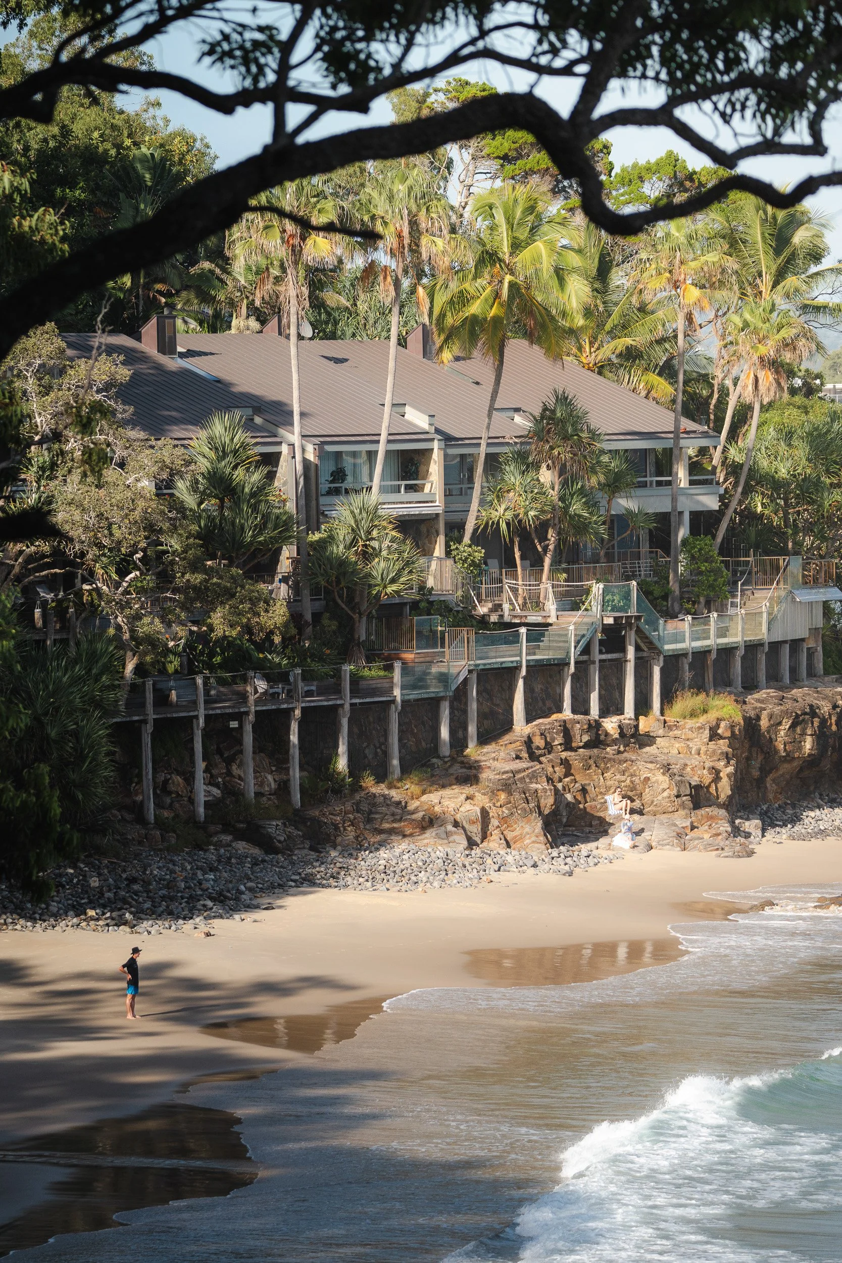 Beachside house with multiple balconies surrounded by palm trees, overlooking a sandy beach with two people near the shore and a rocky cliff.