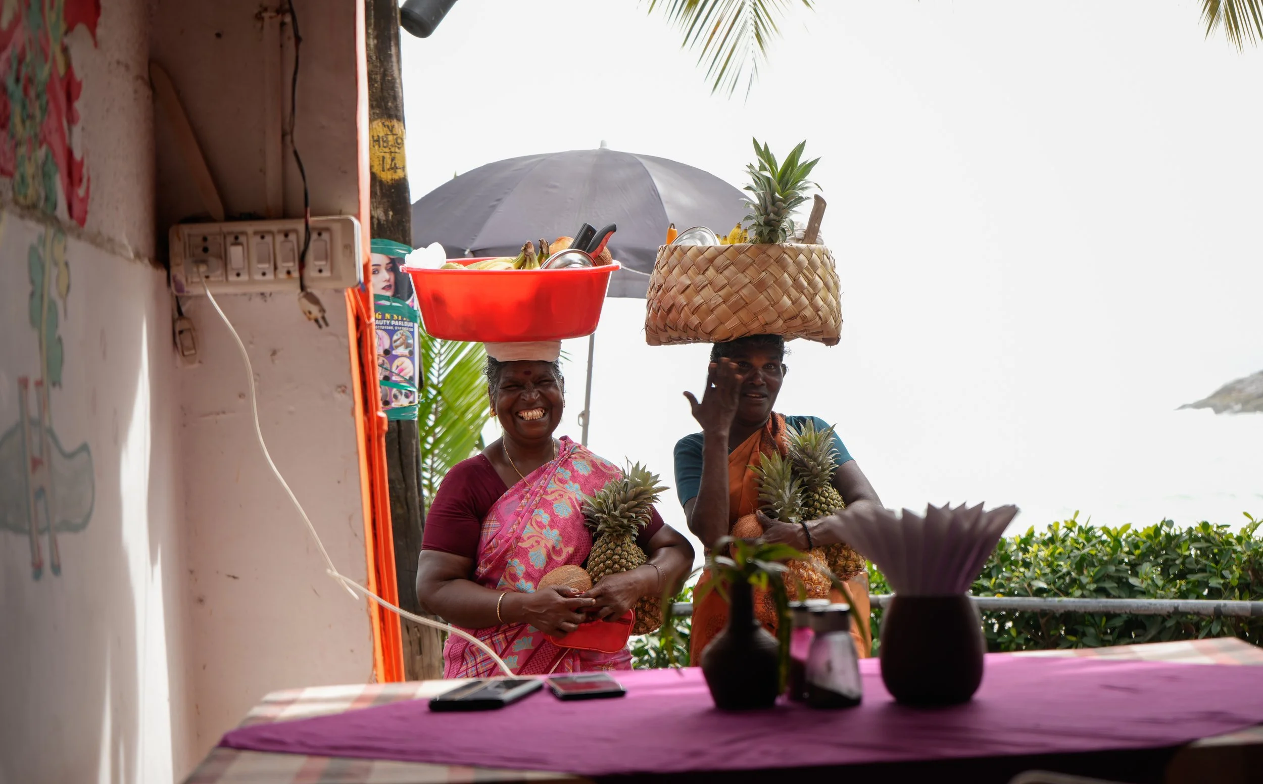 Lifestyle travel photography of two women in vibrant saris carrying tropical fruit by the sea