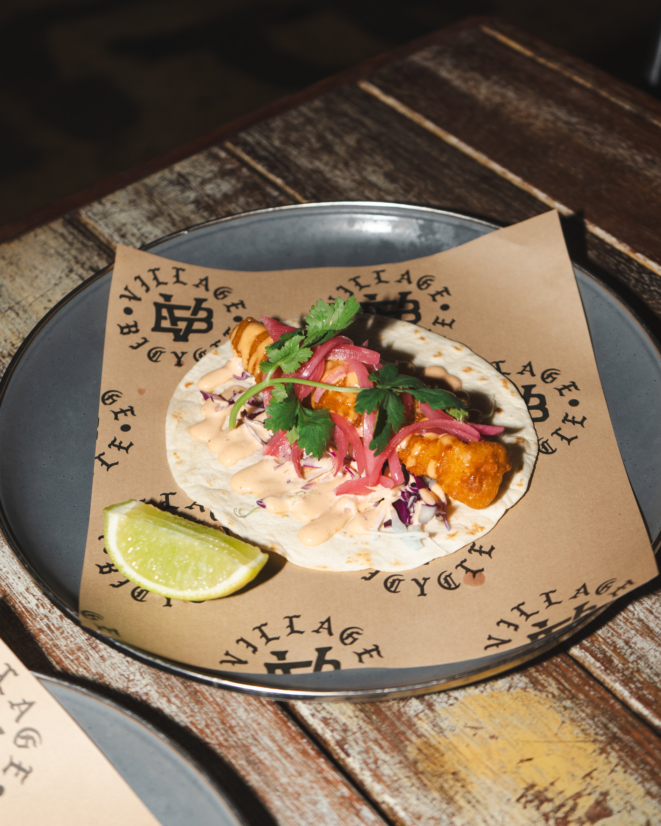 Fish taco with cilantro, pickled red onions, and sauce on a plate with lime wedge, on a wooden table.