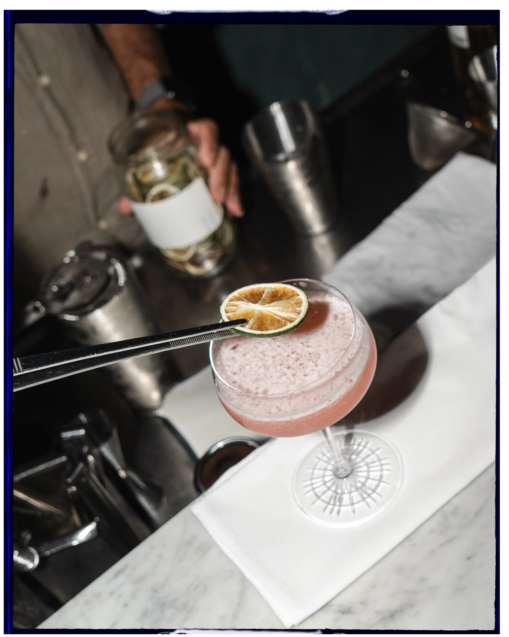 A pink cocktail garnished with a dried lime slice and a black straw, placed on a white napkin on a marble bar counter.