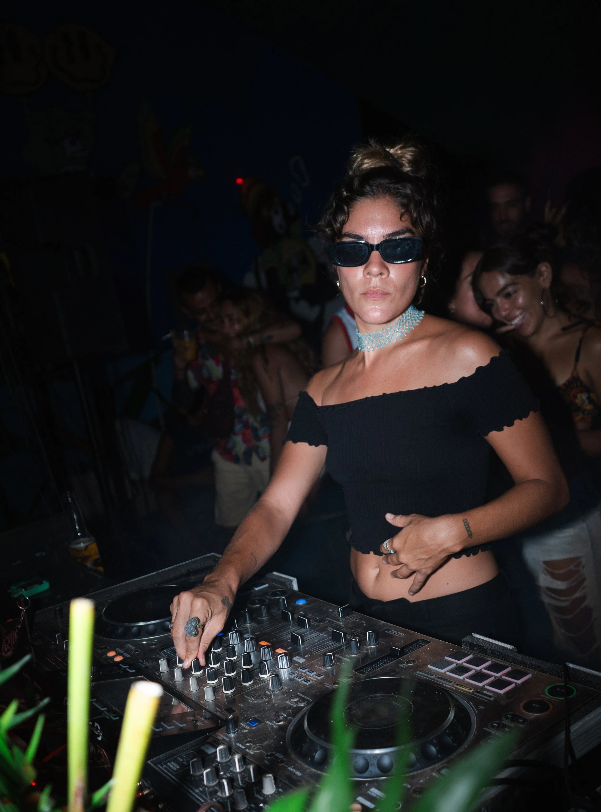 Female DJ wearing sunglasses and black off shoulder top performing on a DJ mixer at a crowded club, high energy atmosphere, event and nightlife photography