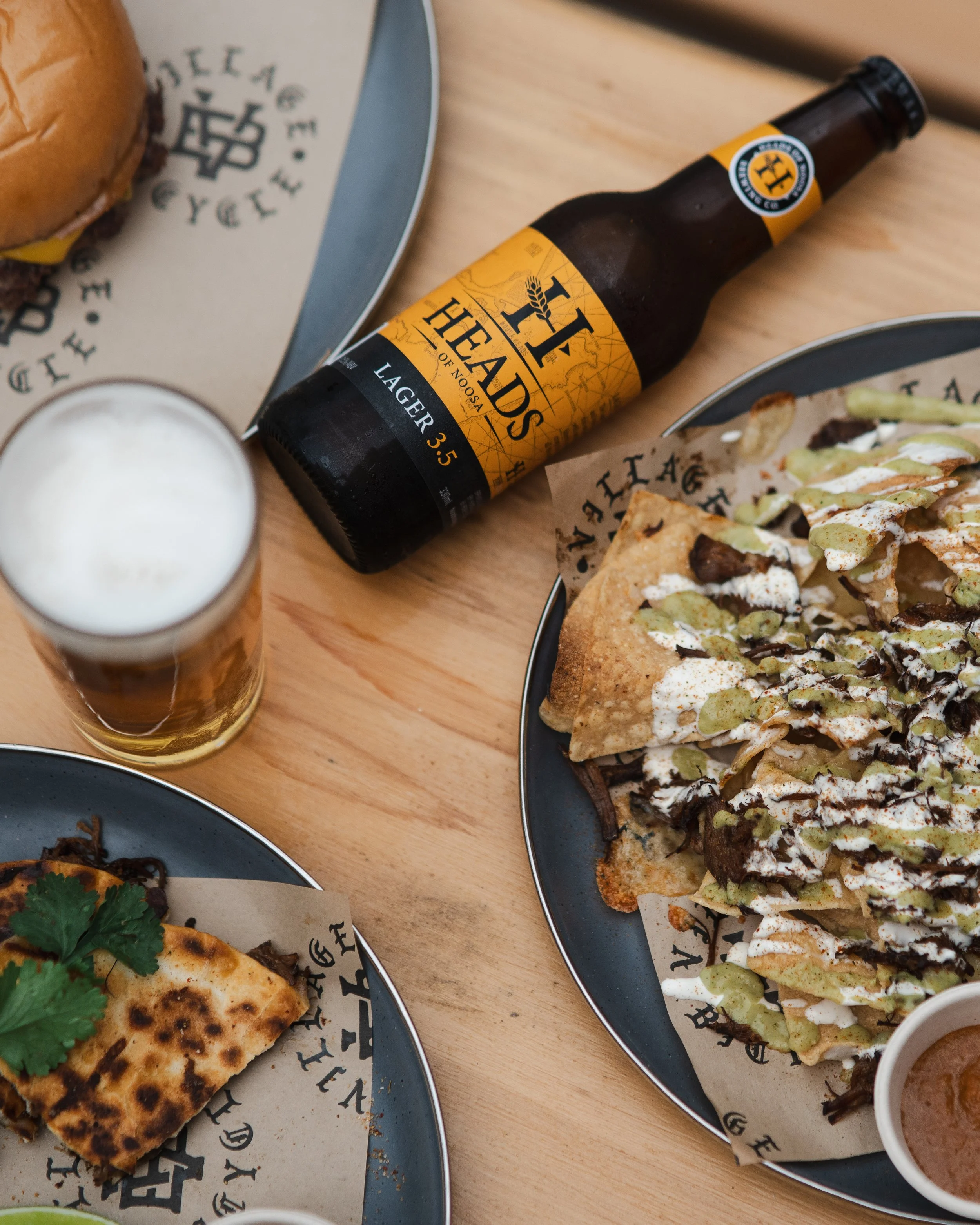 A table with plates of loaded nachos, a cheeseburger, a slice of pizza, a bottle of Toad the Wet Sprocket beer, and a glass of beer.