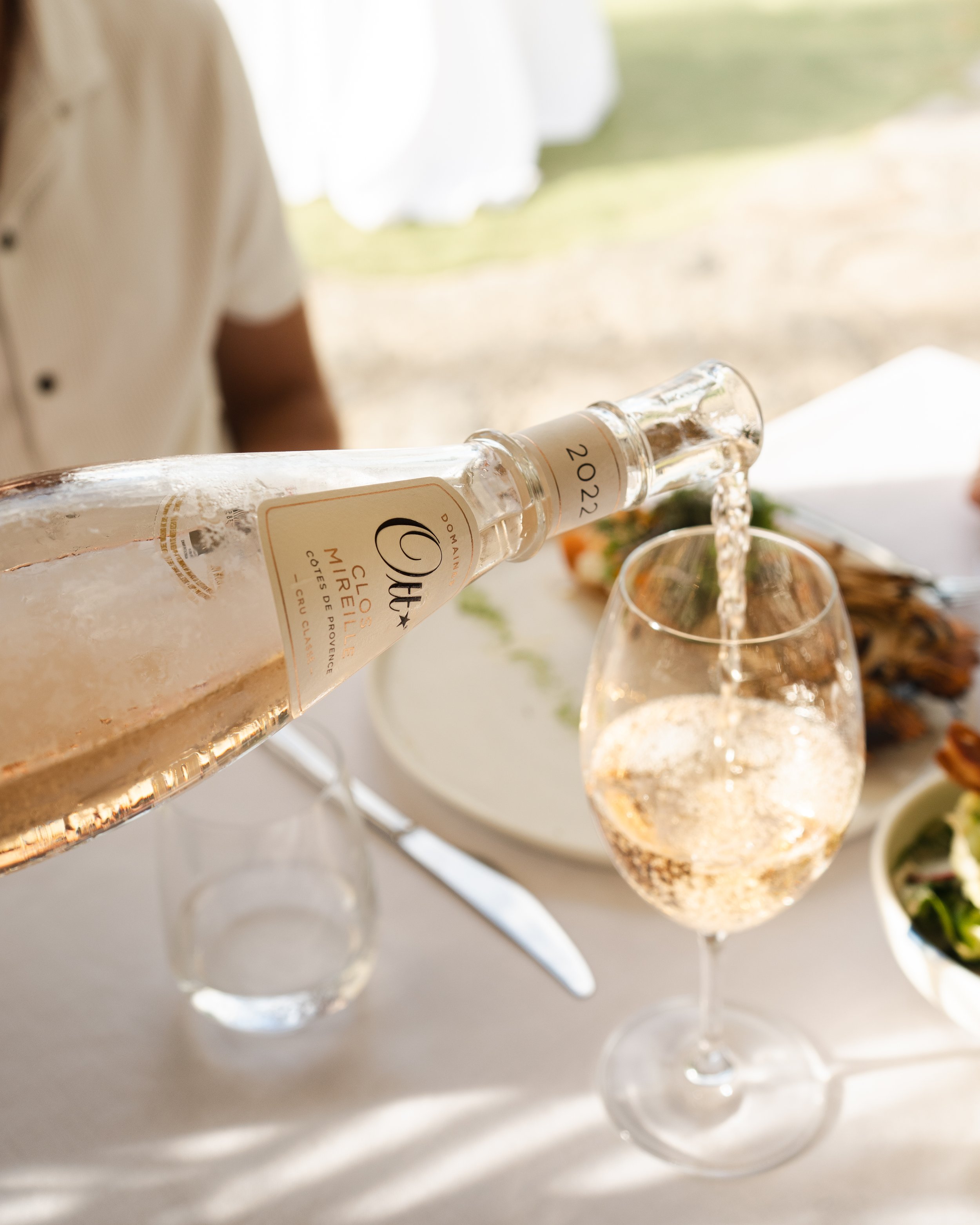 A person pours rosé wine from a bottle into a wine glass on a table set for a meal with plates and cutlery, outdoors with sunlight.