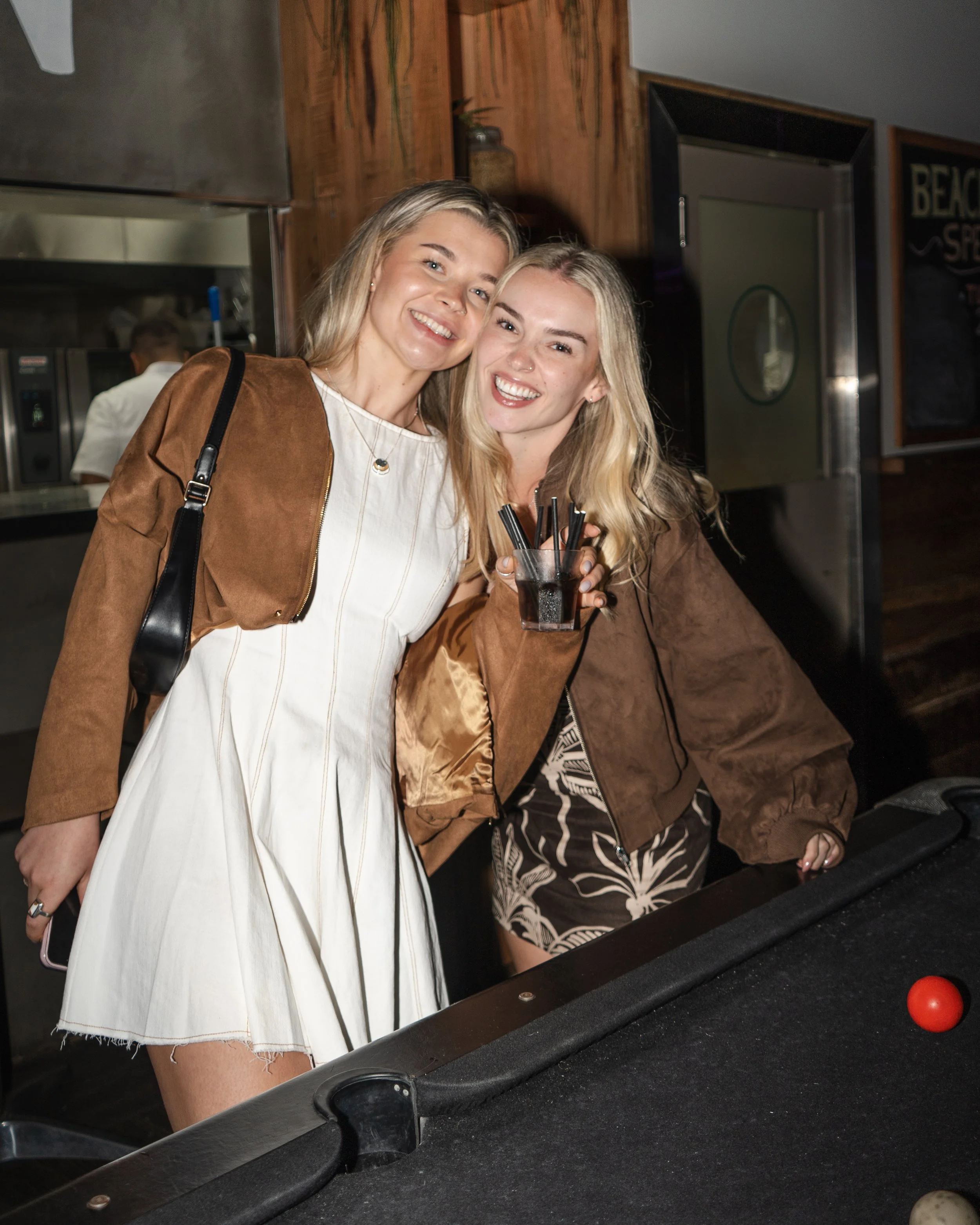Two women posing with drinks next to a pool table in a Noosa bar, casual outfits and lively setting, Sunshine Coast event and nightlife photography