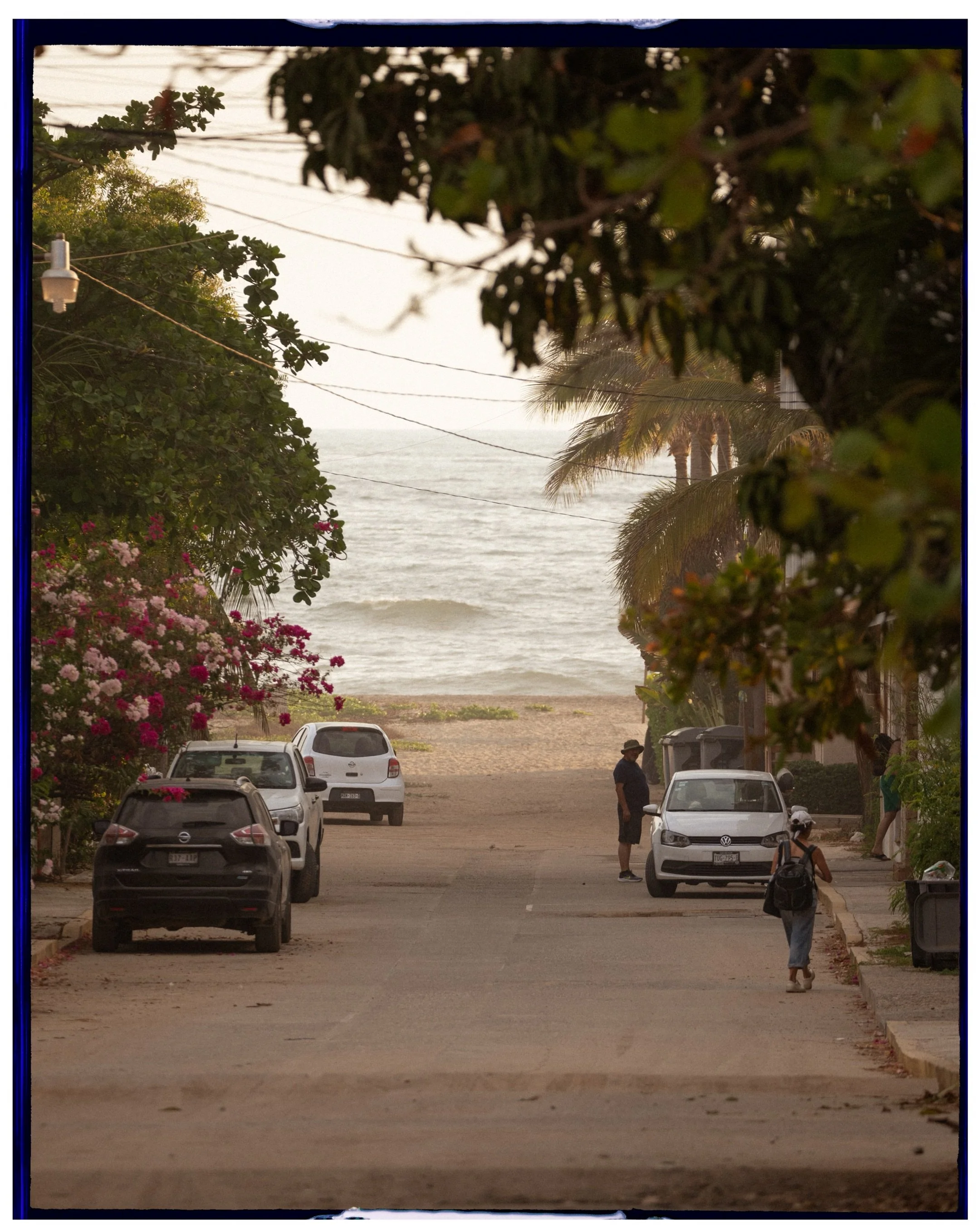 Cinematic travel photography of a scenic street leading to a tropical beach with lush greenery and parked cars