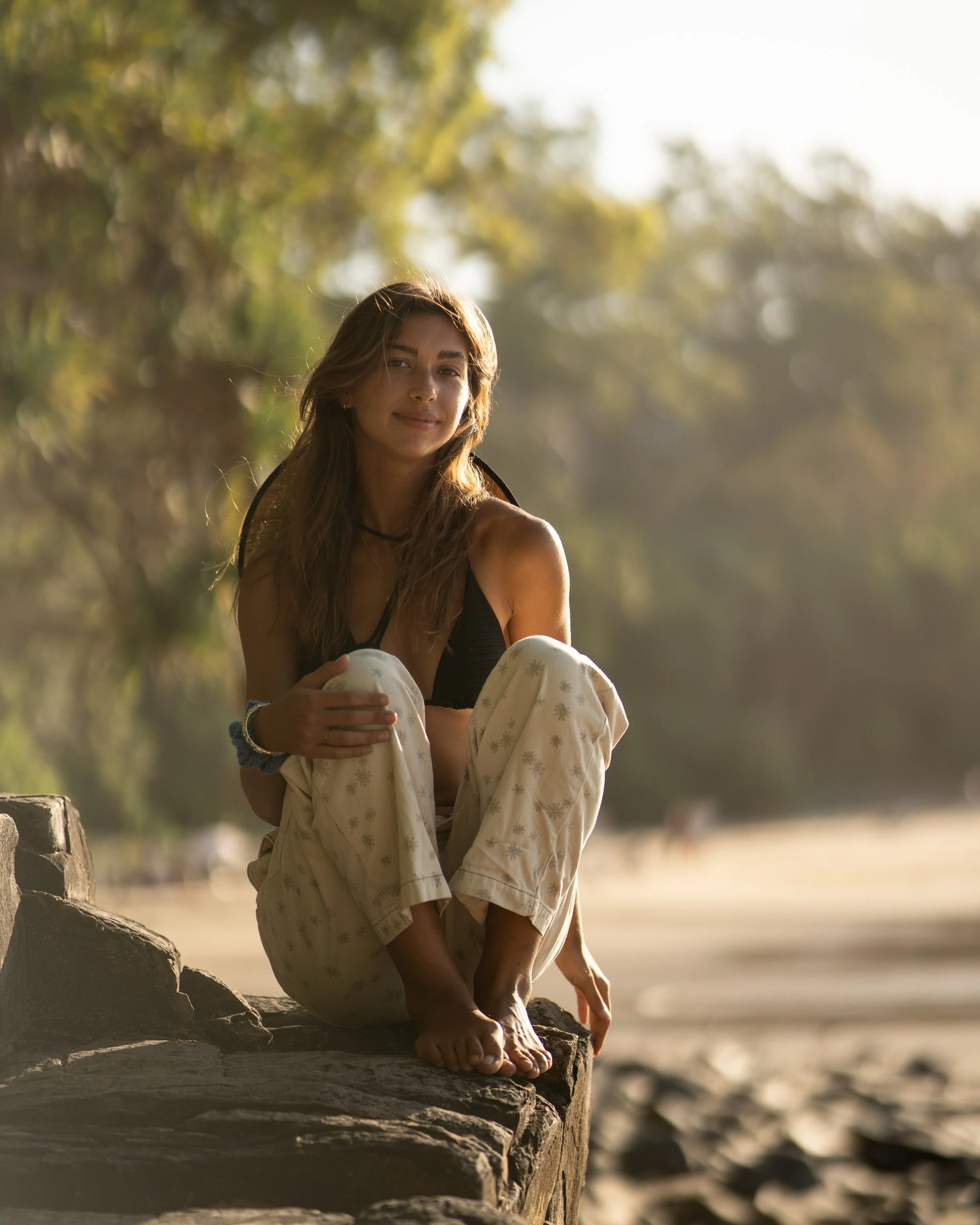 A young woman with long hair sitting with her knees up on a large rock on the beach, wearing a black bikini top and light-colored patterned pants, with trees in the background and sunlight highlighting her face.