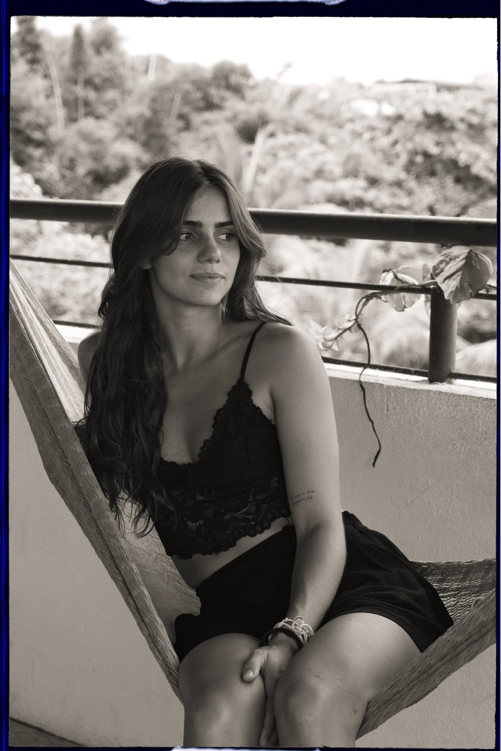 A young woman with long dark hair sitting in a hammock on a balcony, wearing a black lace tank top and shorts, looking pensively to the side, with a lush, green outdoor landscape in the background.