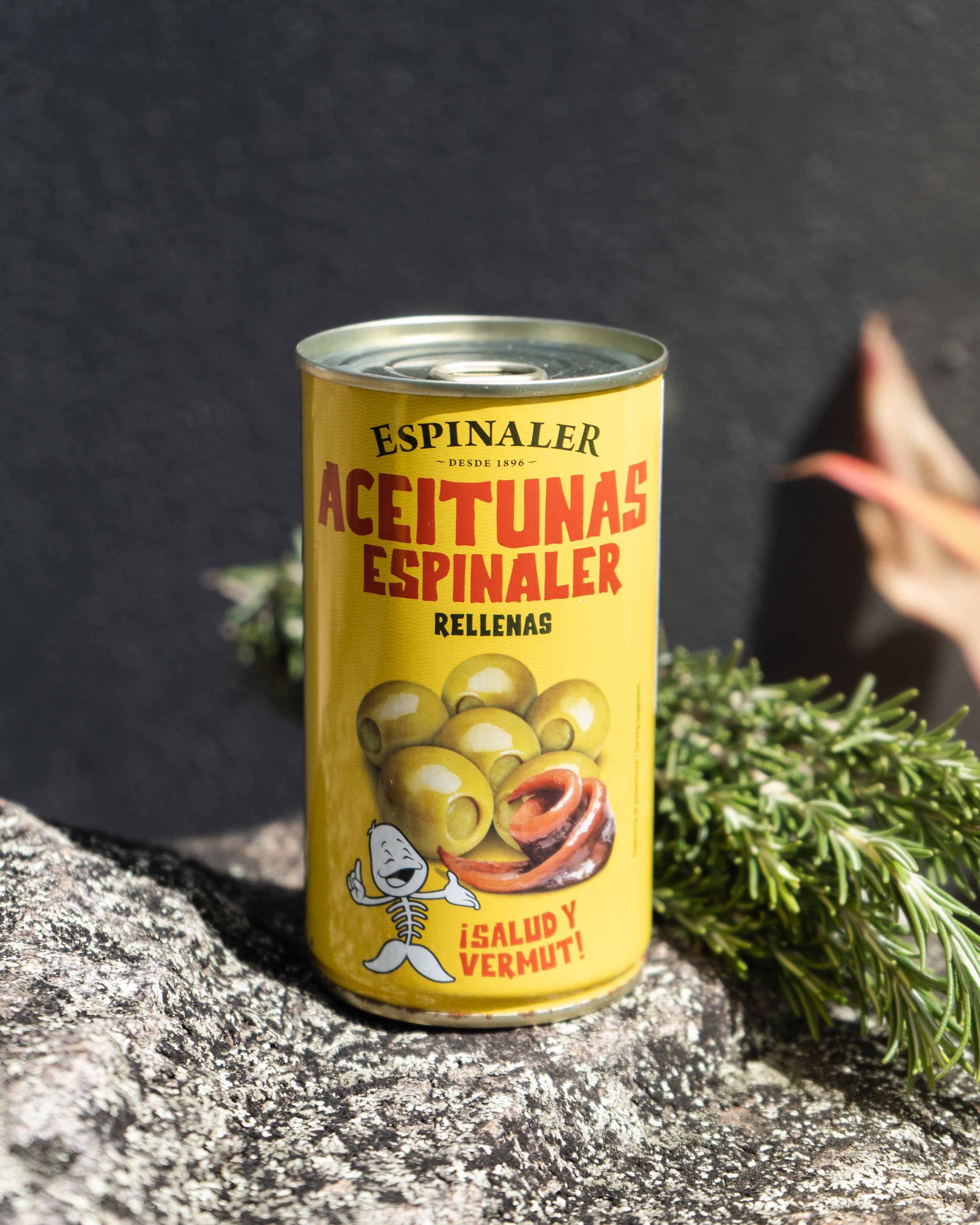 Product photography of a Yellow can of Espinaler brand stuffed green olives, placed outdoors on a rock with some greenery in the background.