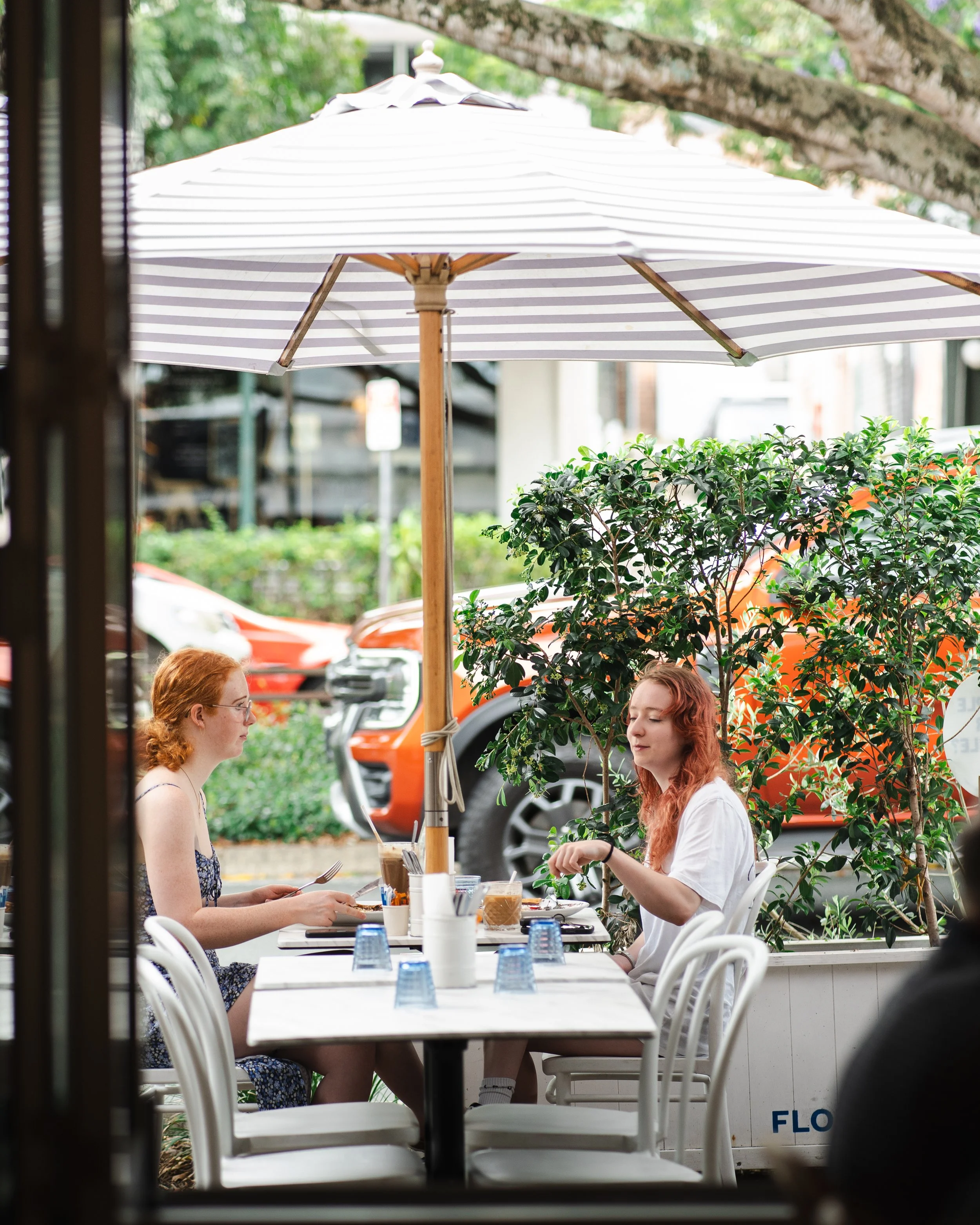Two women sit at a table outside a cafe under a large striped umbrella, surrounded by greenery, parked cars, and trees.