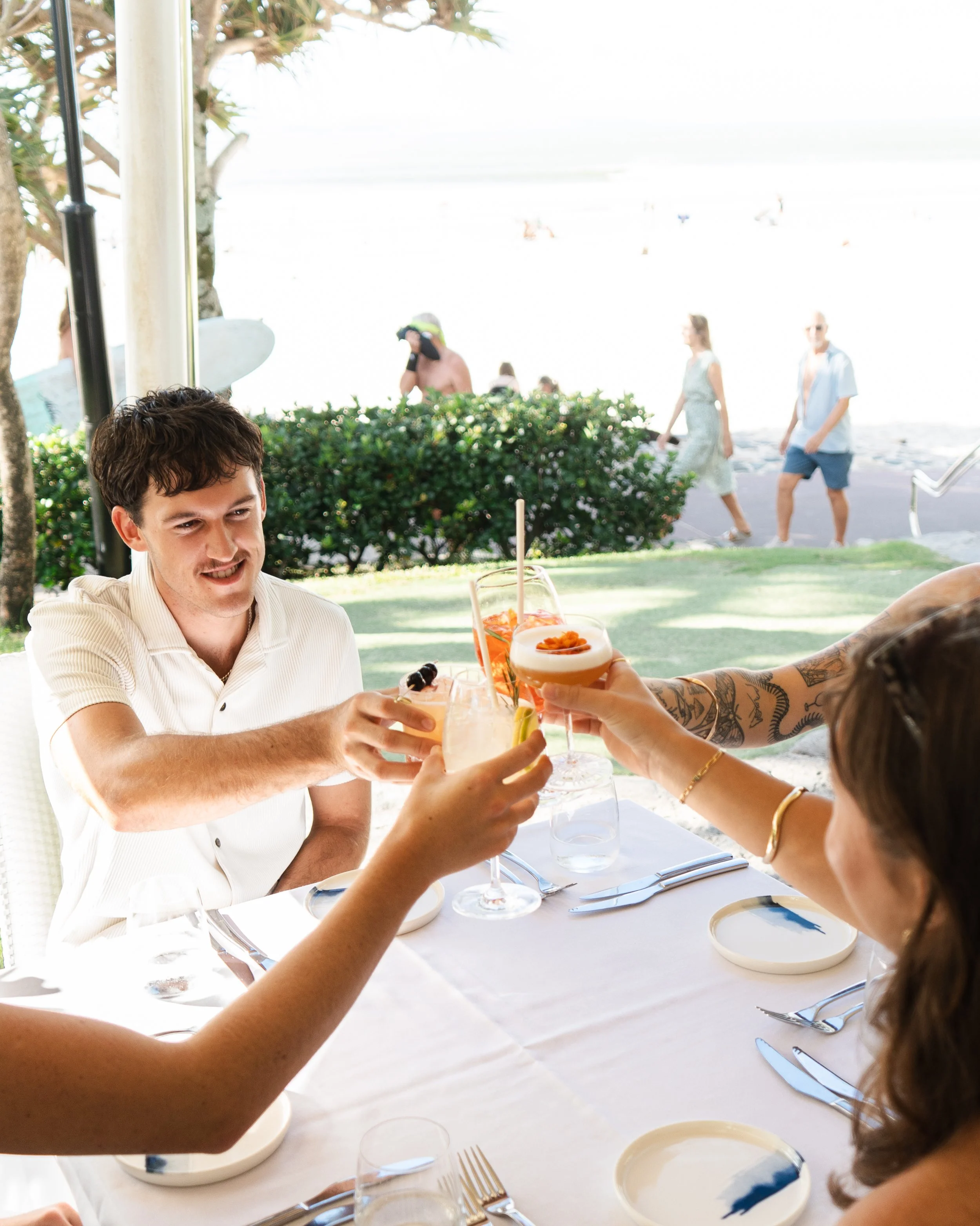 People at a beachside restaurant raising glasses for a toast, with the beach and beachgoers in the background.