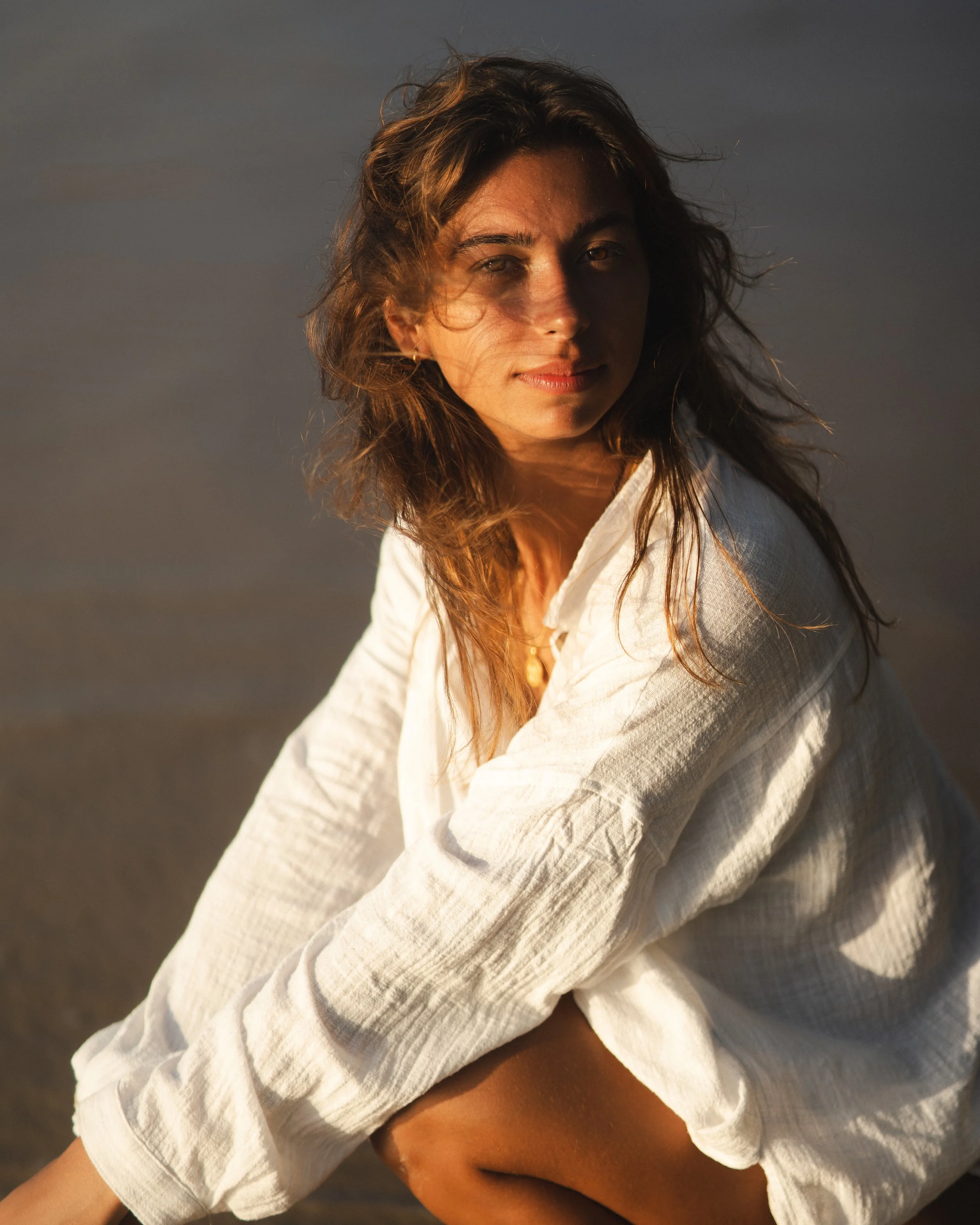 Young woman with brown hair in a white shirt sitting outdoors during sunset or sunrise.