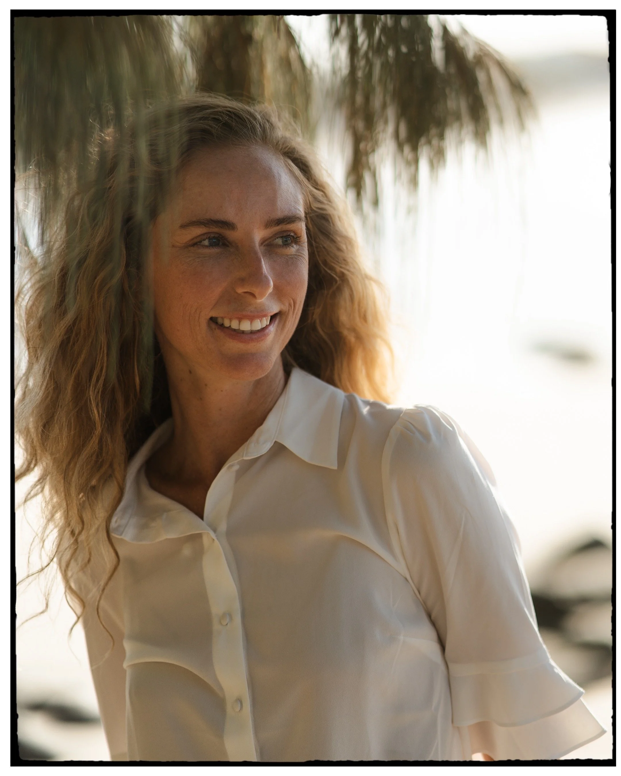 A woman with curly blonde hair smiling, wearing a white blouse, standing outdoors with trees and water in the background.