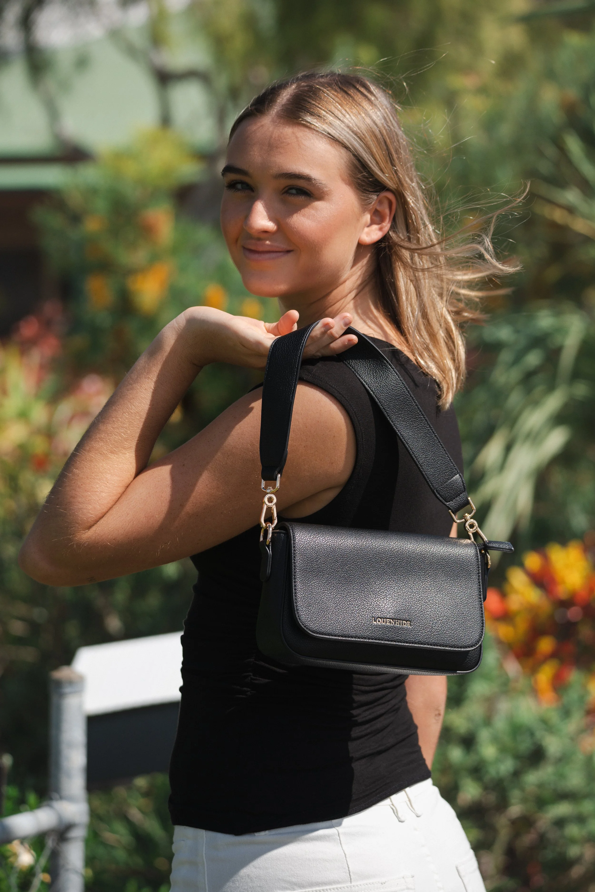 A woman with shoulder-length blonde hair turning towards the camera outdoors, holding a black handbag with a strap over her shoulder, in front of a background of green foliage.