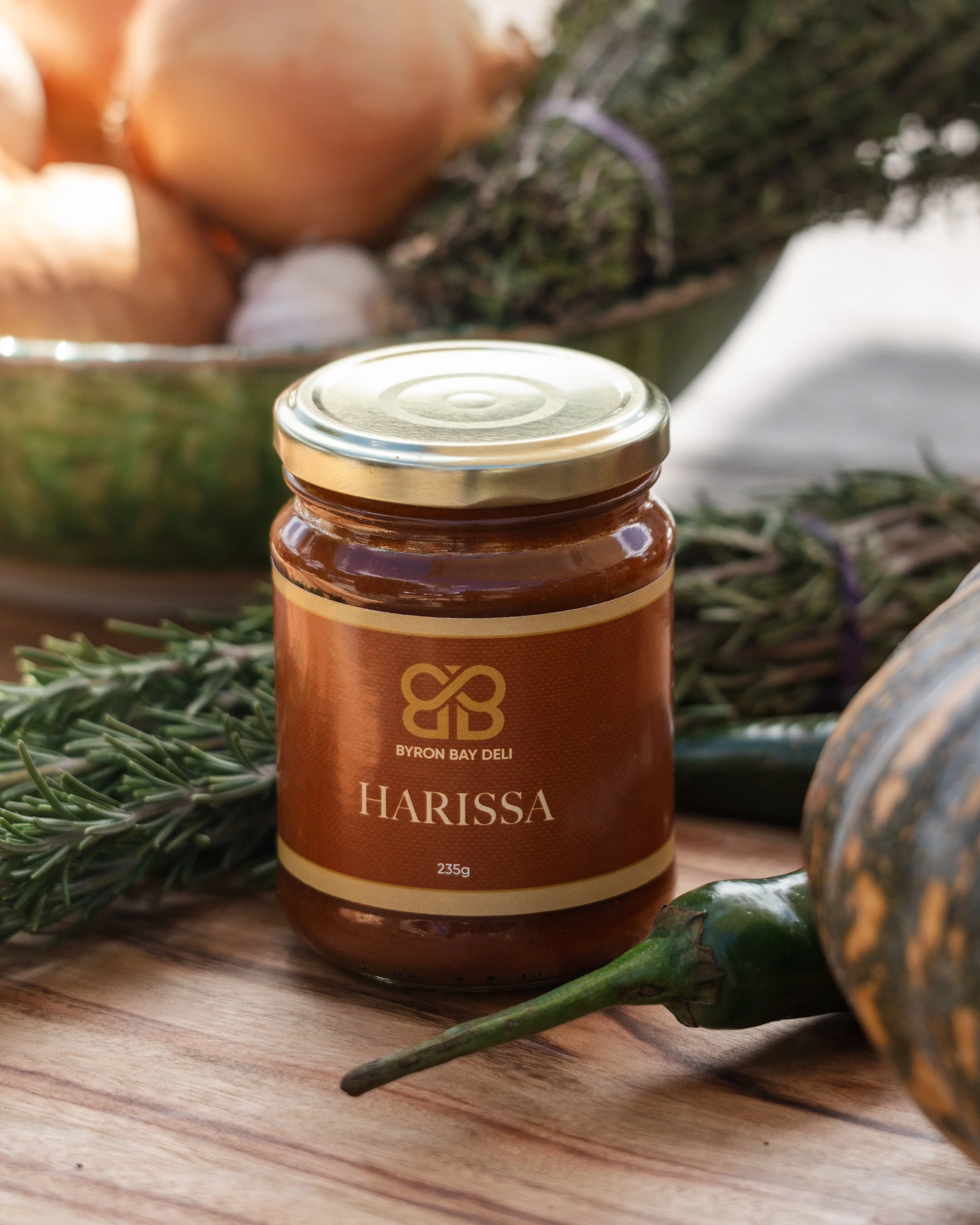 Product photography of a jar labeled "Harissa" from Byron Bay Deli in the foreground with a sprig of rosemary, a green chili, and an onion in the background on a wooden surface.