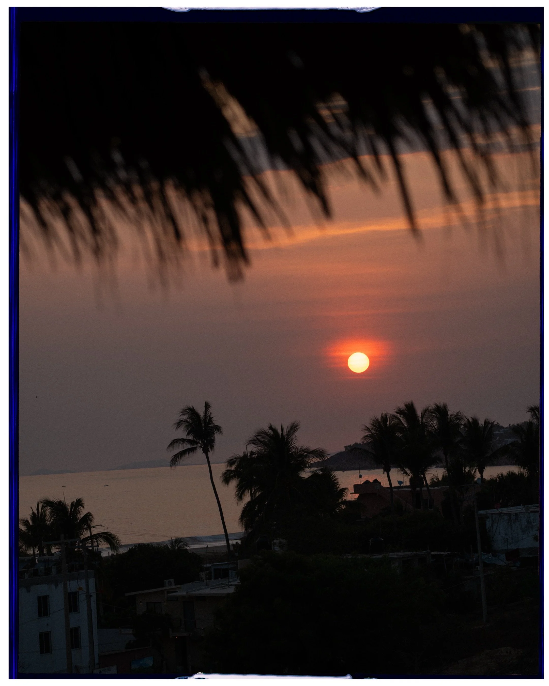 Editorial destination photography of a tropical sunset over palm trees and coastal buildings, framed by a thatched roof