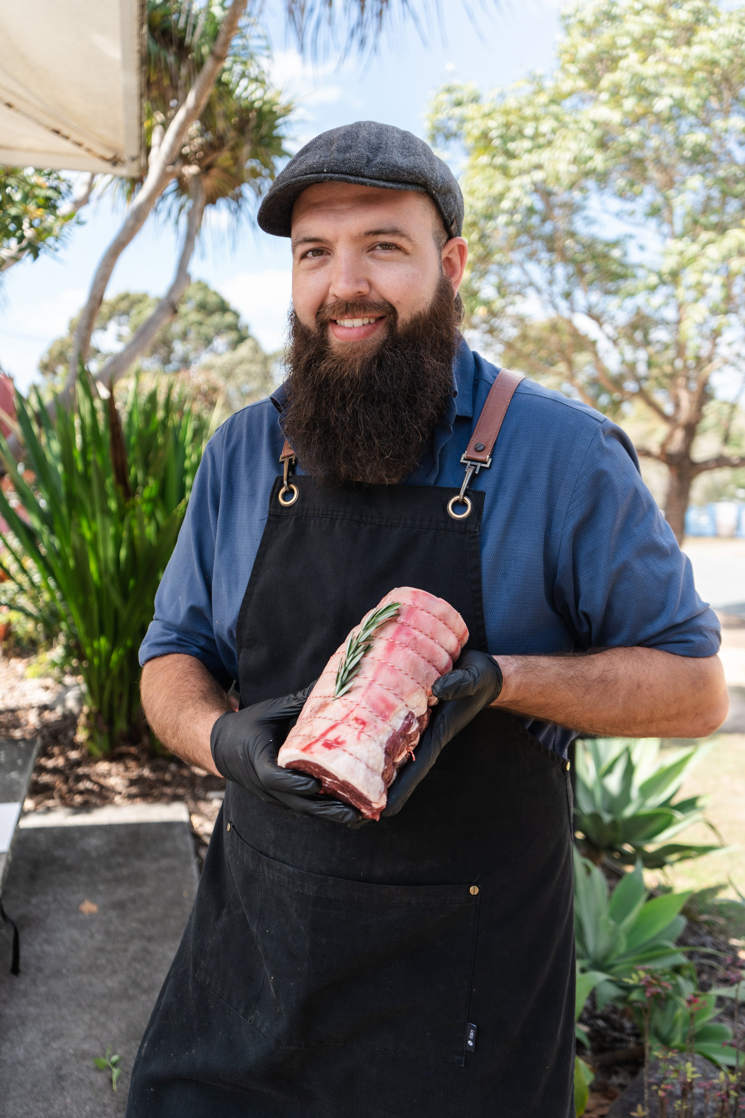 Man with a beard and hat smiling while holding a raw piece of meat with a sprig of rosemary, outdoors on a sunny day.