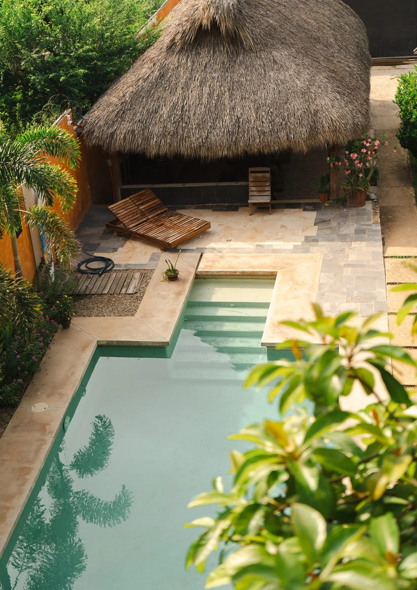 A backyard with a swimming pool, a thatched roof hut, wooden lounge chair, potted plants, and lush green trees.