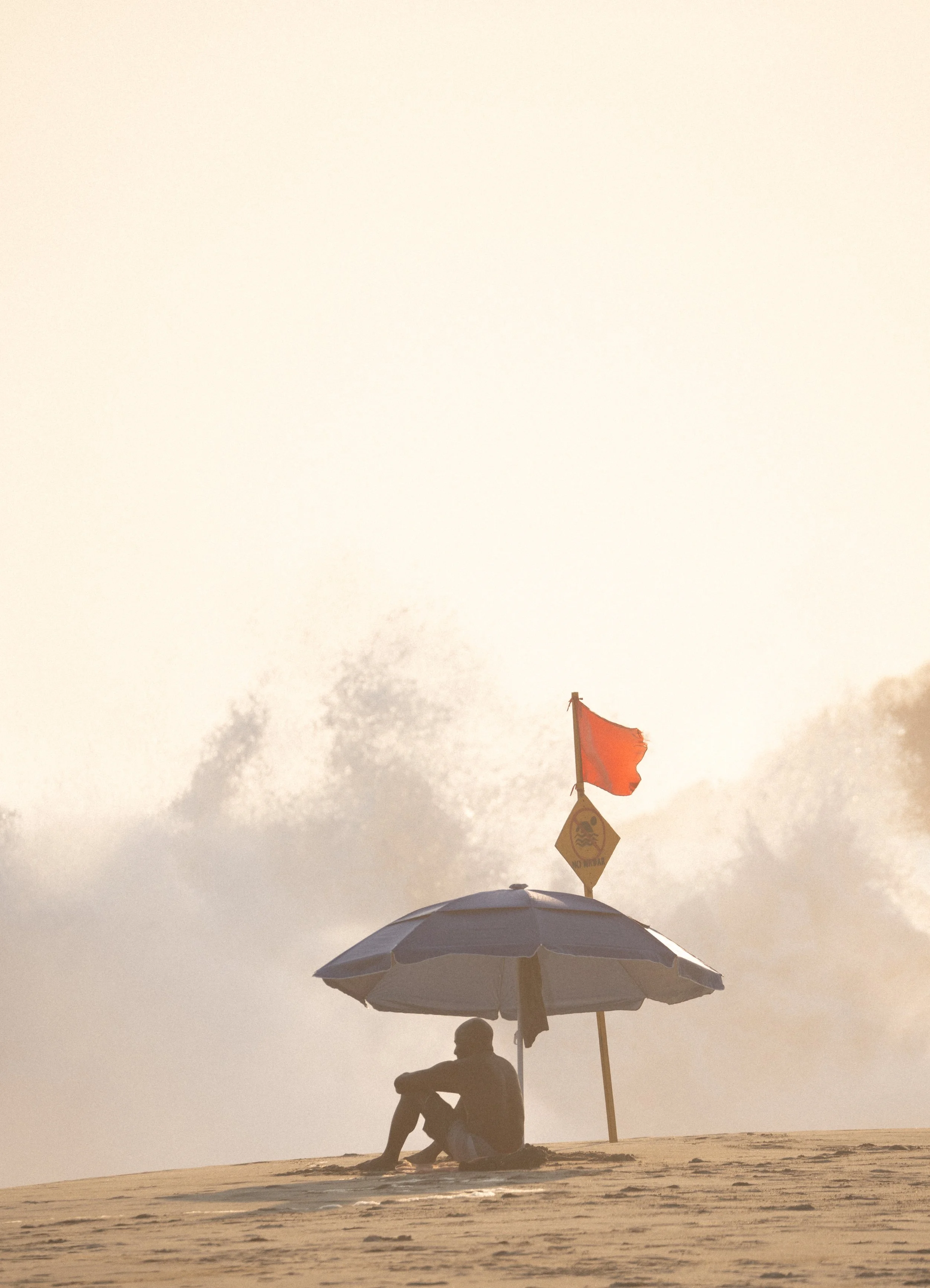 Editorial travel photography of a person under a beach umbrella on a foggy coastal beach with a warning flag