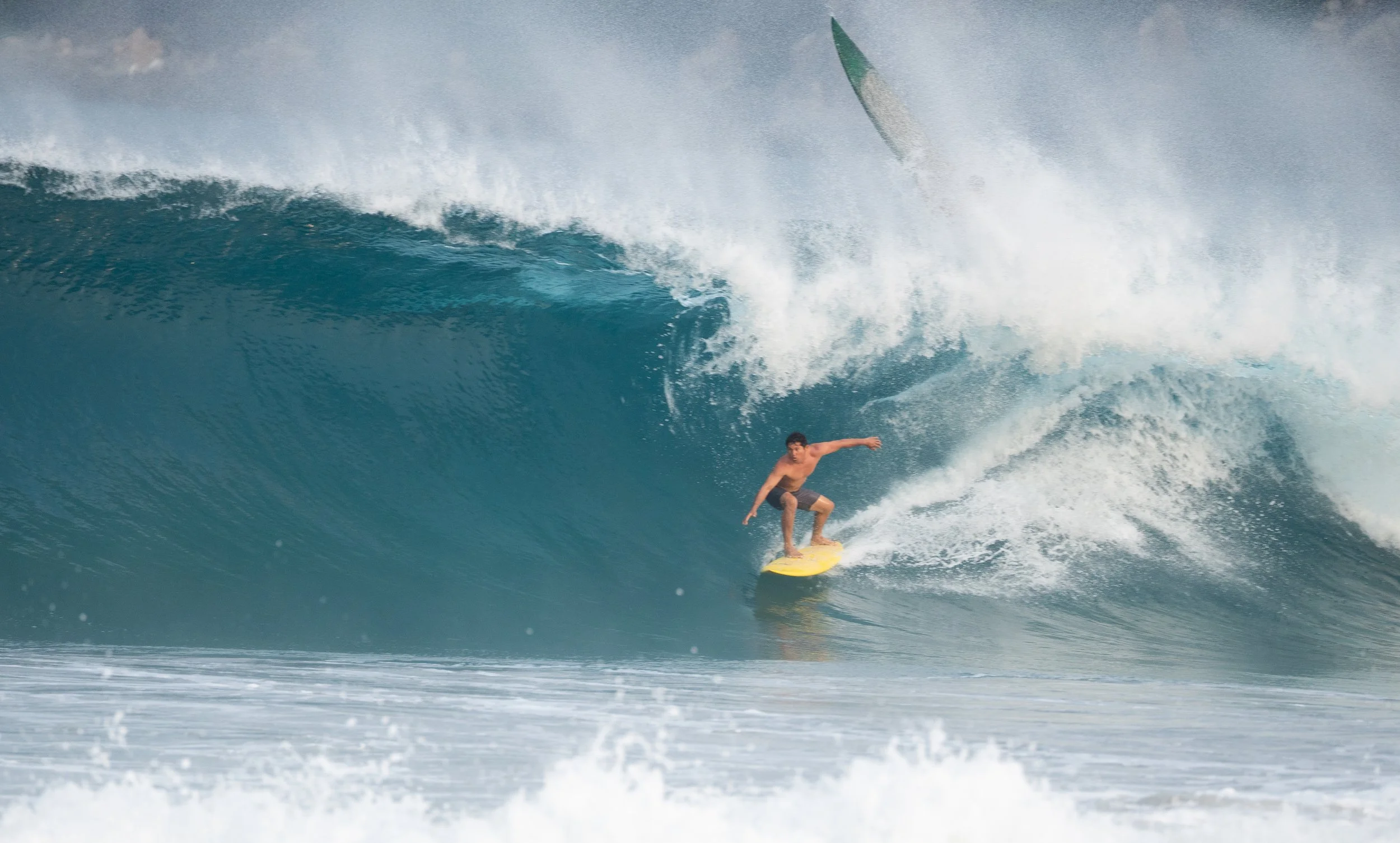 A male surfer riding inside a large, breaking ocean wave, with a surfboard and a boat in the background.