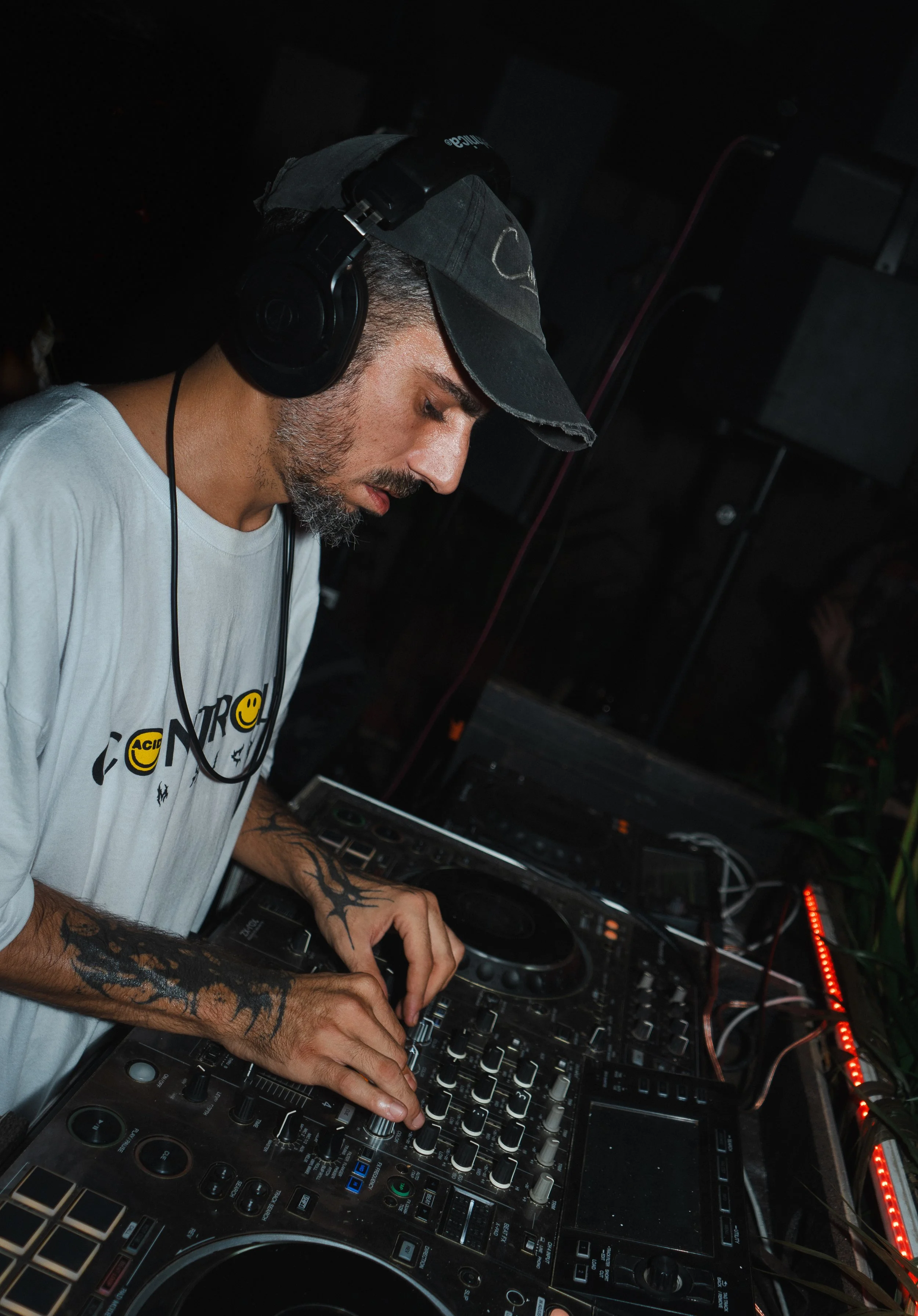 Nightlife photography of a DJ with tattoos on his arms, wearing a black cap and headphones, is focused on mixing music with a professional DJ controller at a dark indoor event
