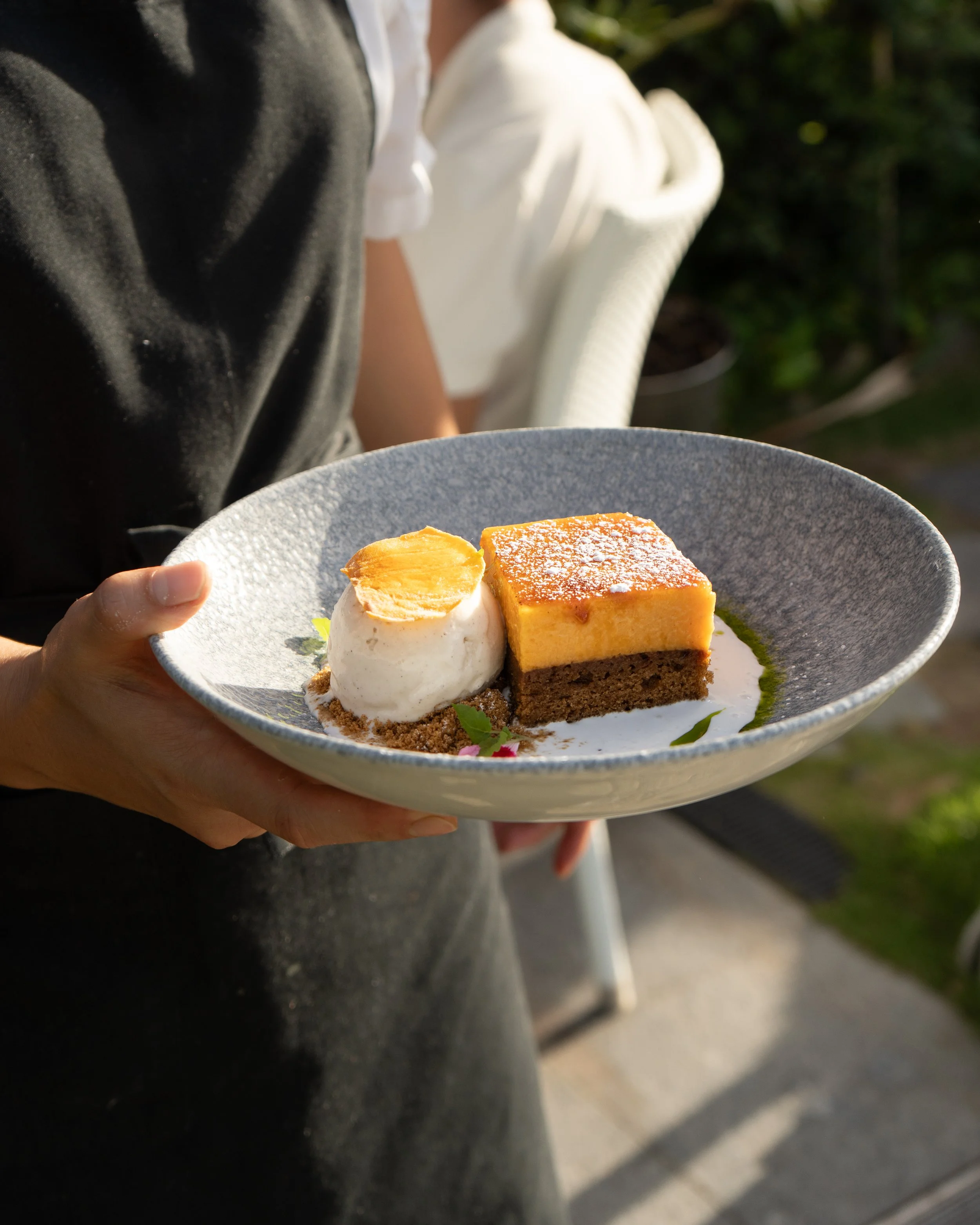 A person holding a gray plate with a slice of cheesecake, a scoop of ice cream topped with a cookie, and garnishes, outdoors with sunlight.