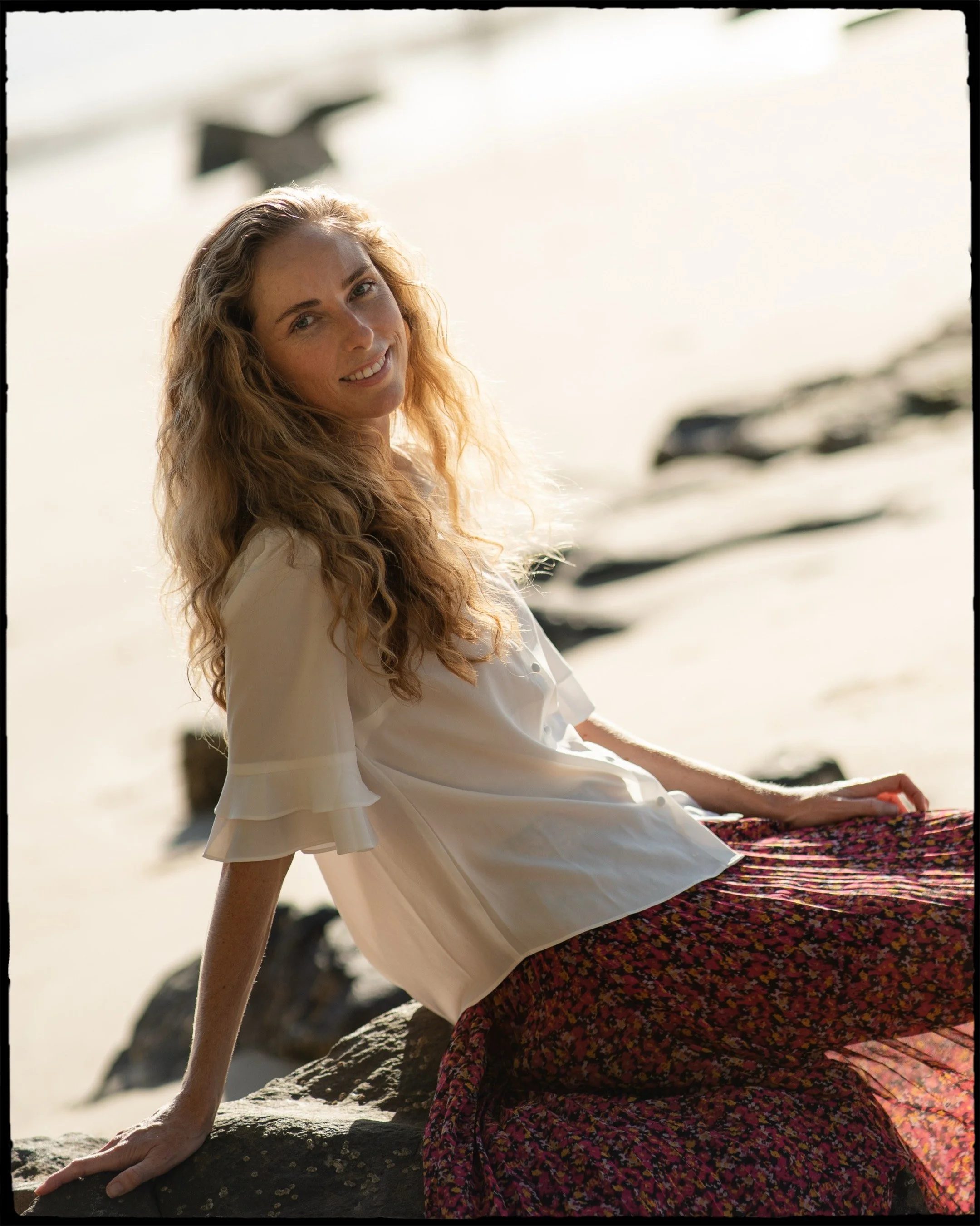 A woman with long, wavy blonde hair sitting on rocks on a beach, smiling at the camera, wearing a white blouse and a colorful skirt, with the ocean and rocks visible in the background.