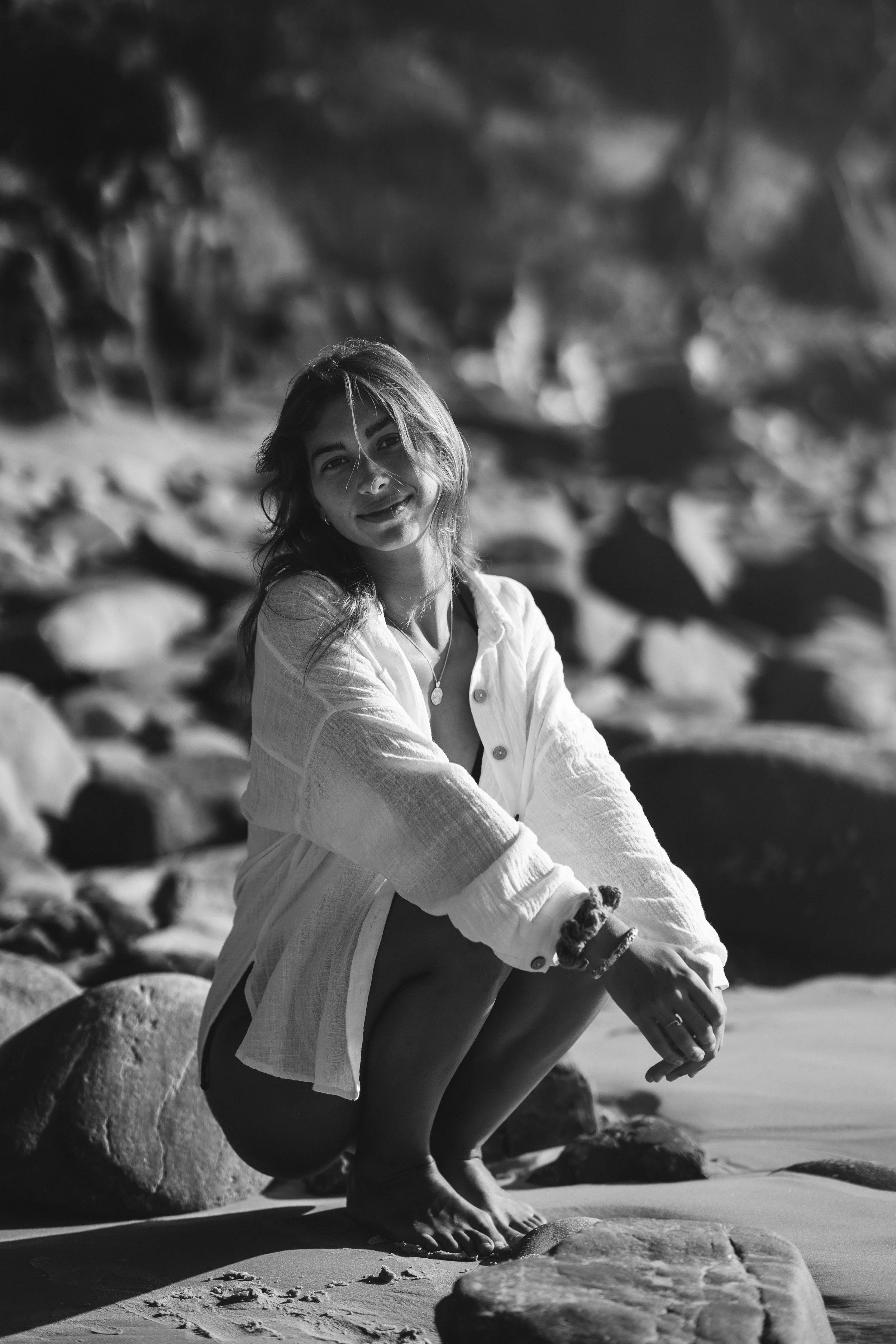 A woman squatting on rocks at the beach, smiling at the camera, in a black and white photo.