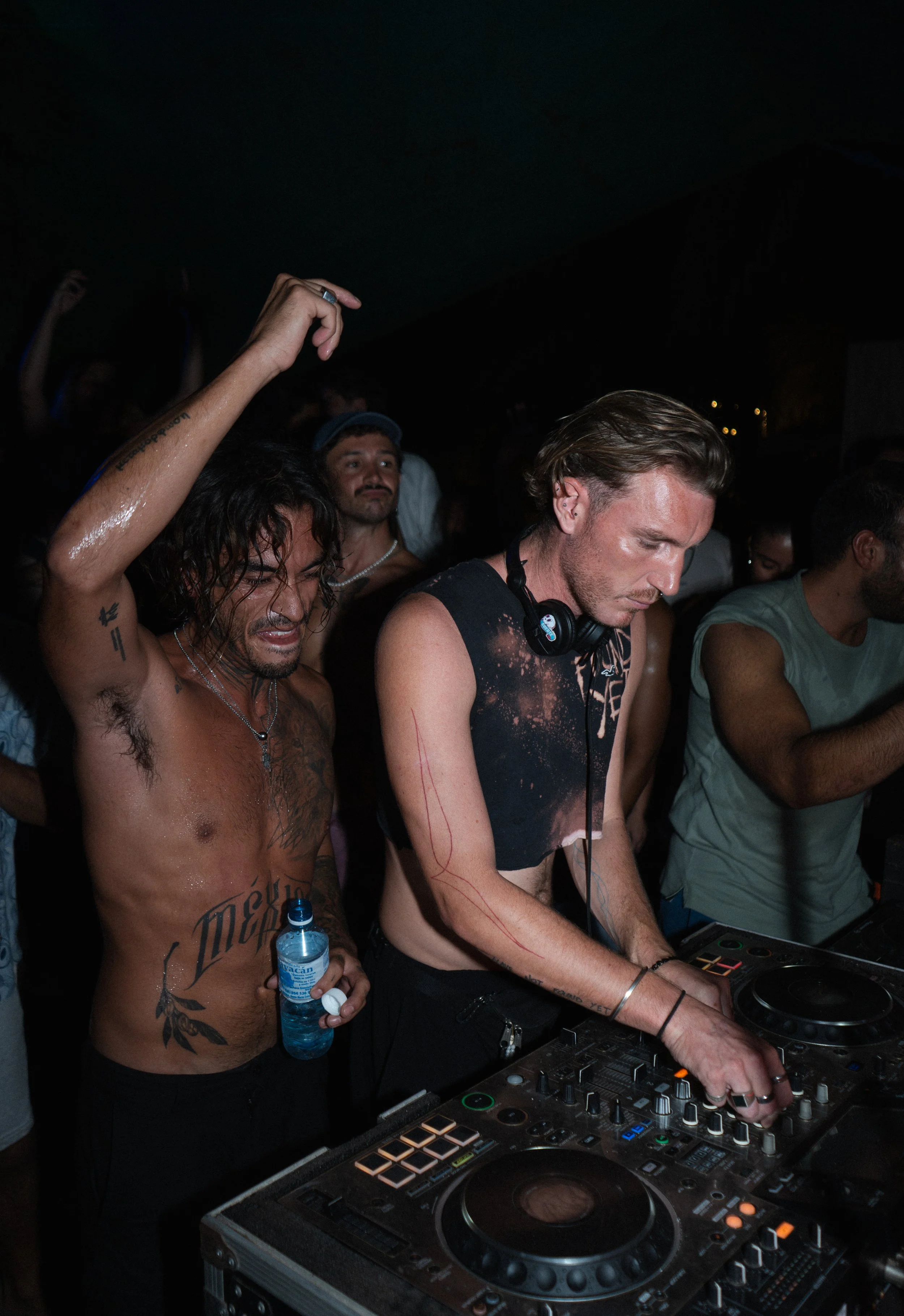 DJ mixing music at a party with dancer holding a water bottle beside him, crowd moving in the background, dynamic event and nightlife photography
