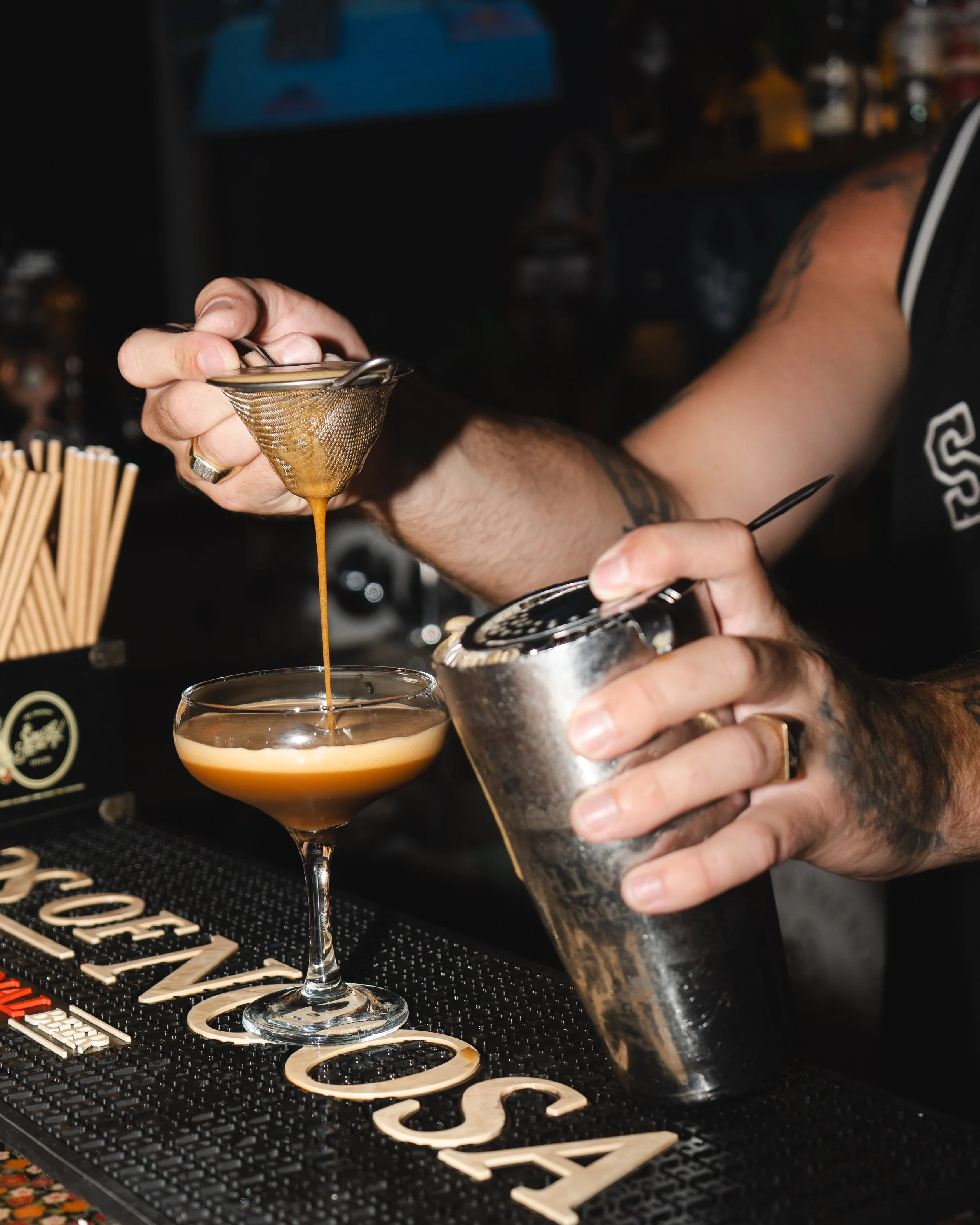 Bartender straining a cocktail into a glass behind a bar counter.