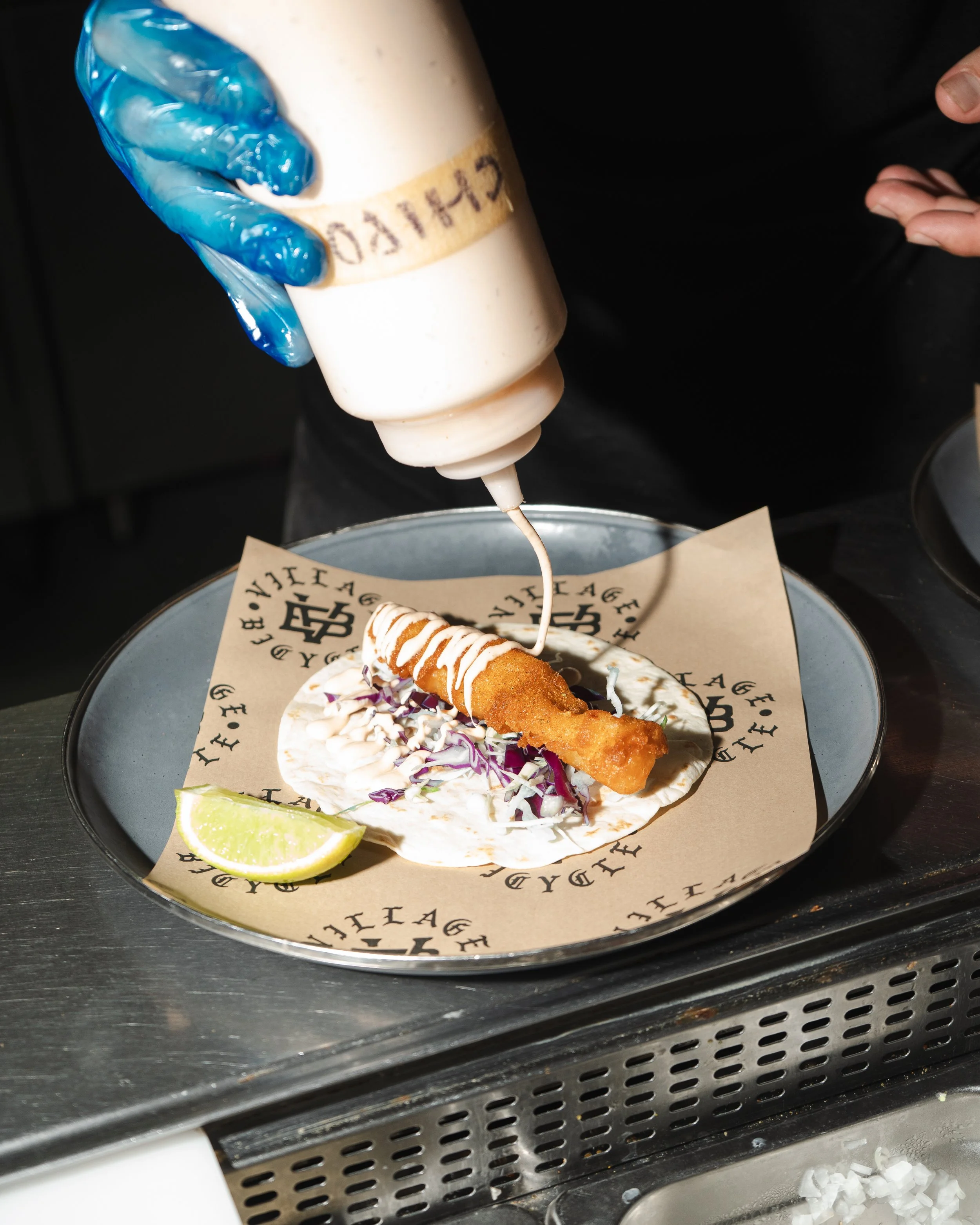 A deep-fried fish taco with shredded purple cabbage, a wedge of lime, and white sauce, topped with a drizzle of white sauce, on a paper-lined plate.