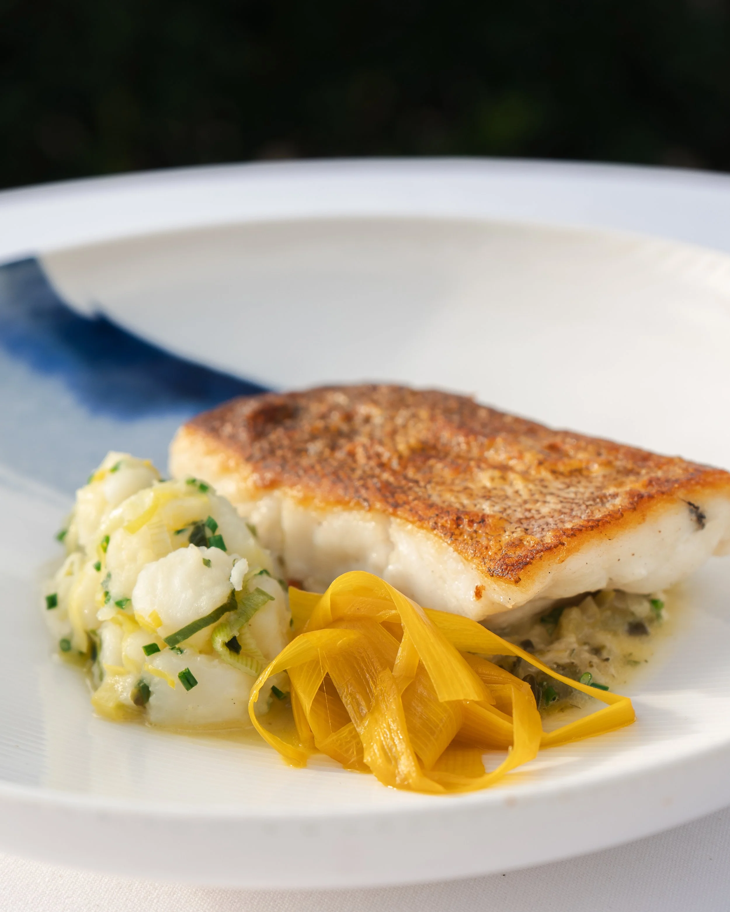 A plate with a cooked fish fillet, mashed potatoes with herbs, and a garnish of yellow pickled vegetables.